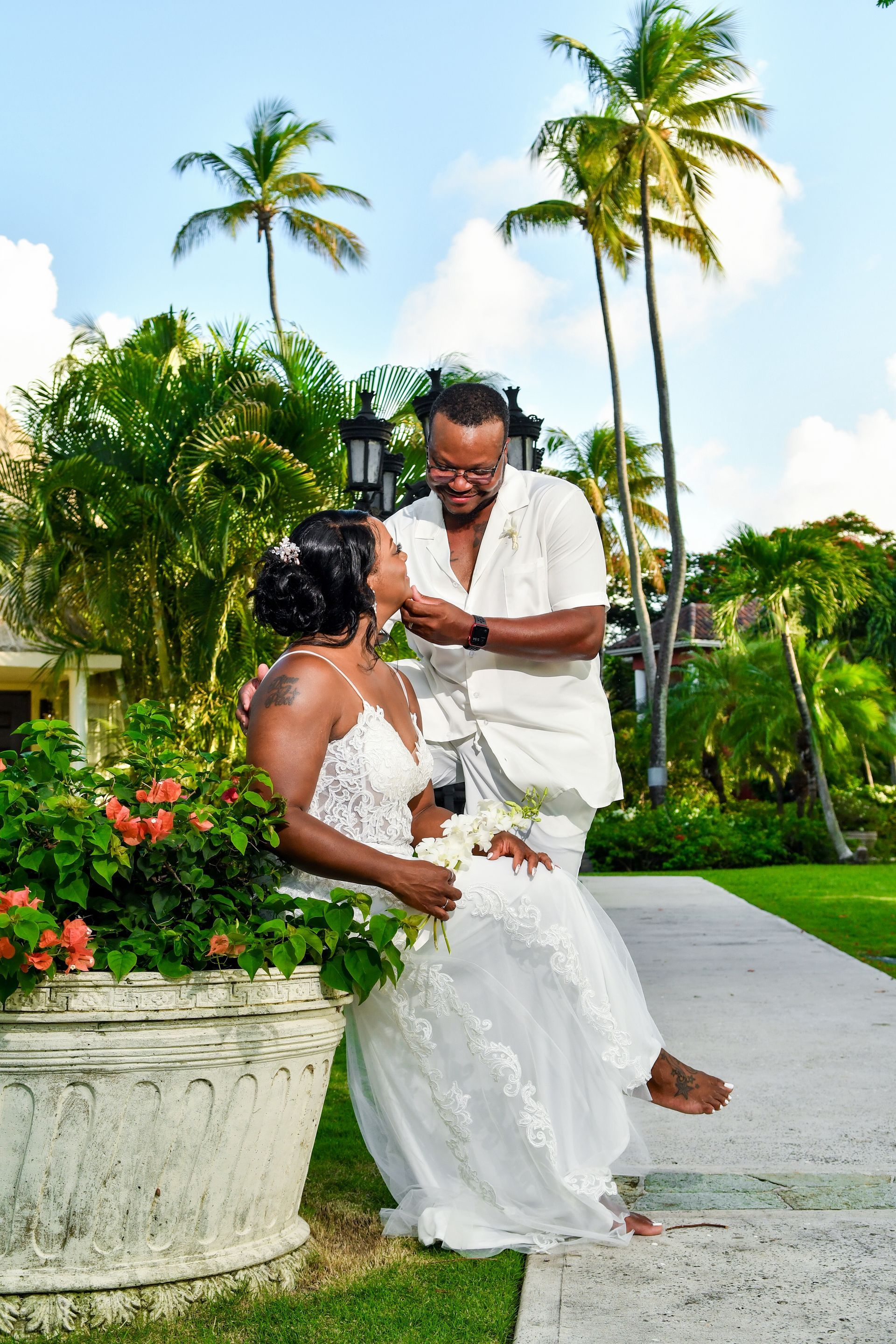 Couple in white on a tropical garden path, with palm trees and lush greenery in the background.