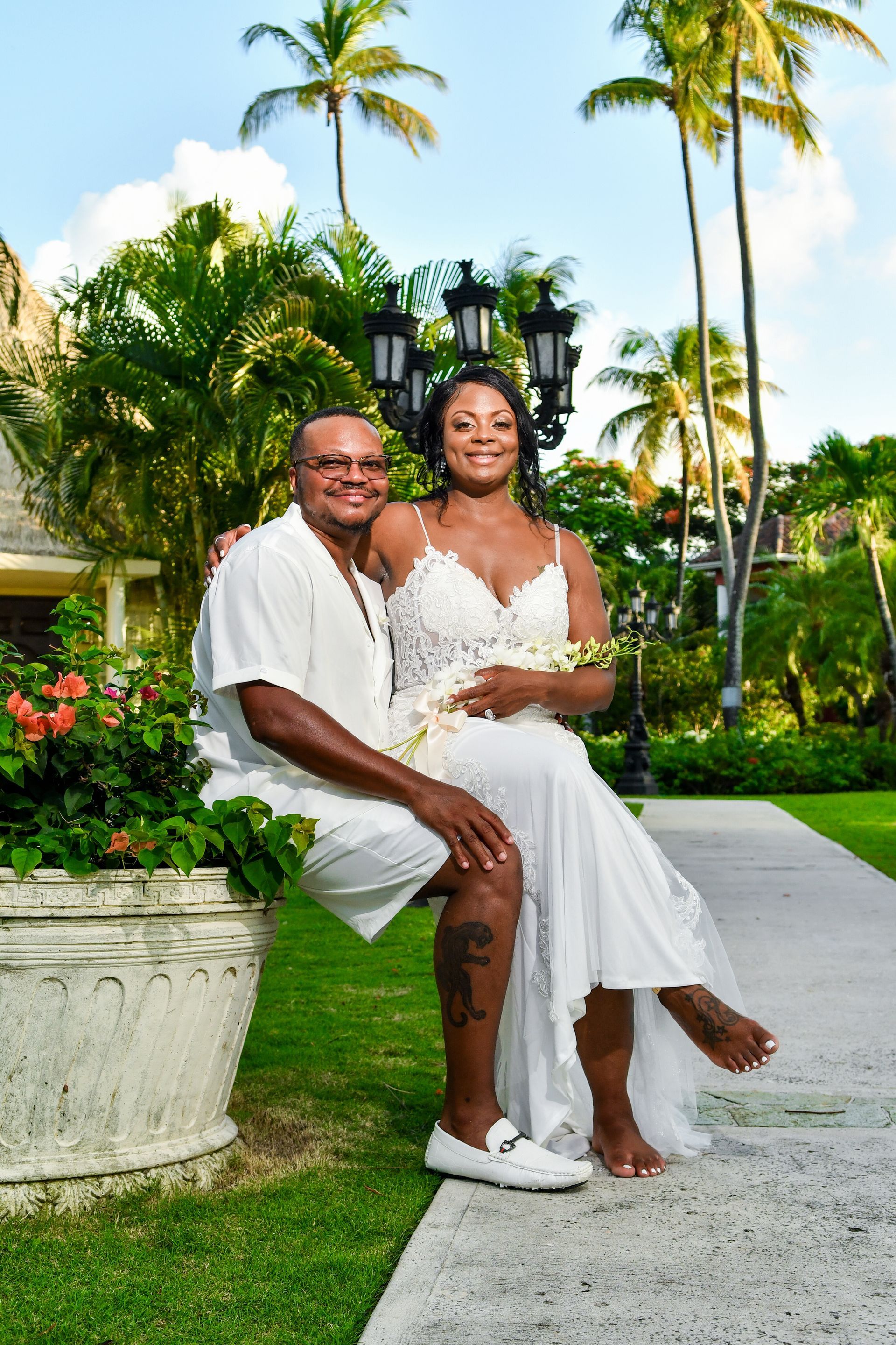 Couple in white outfits posing seated outdoors in a tropical garden with palm trees and flowers