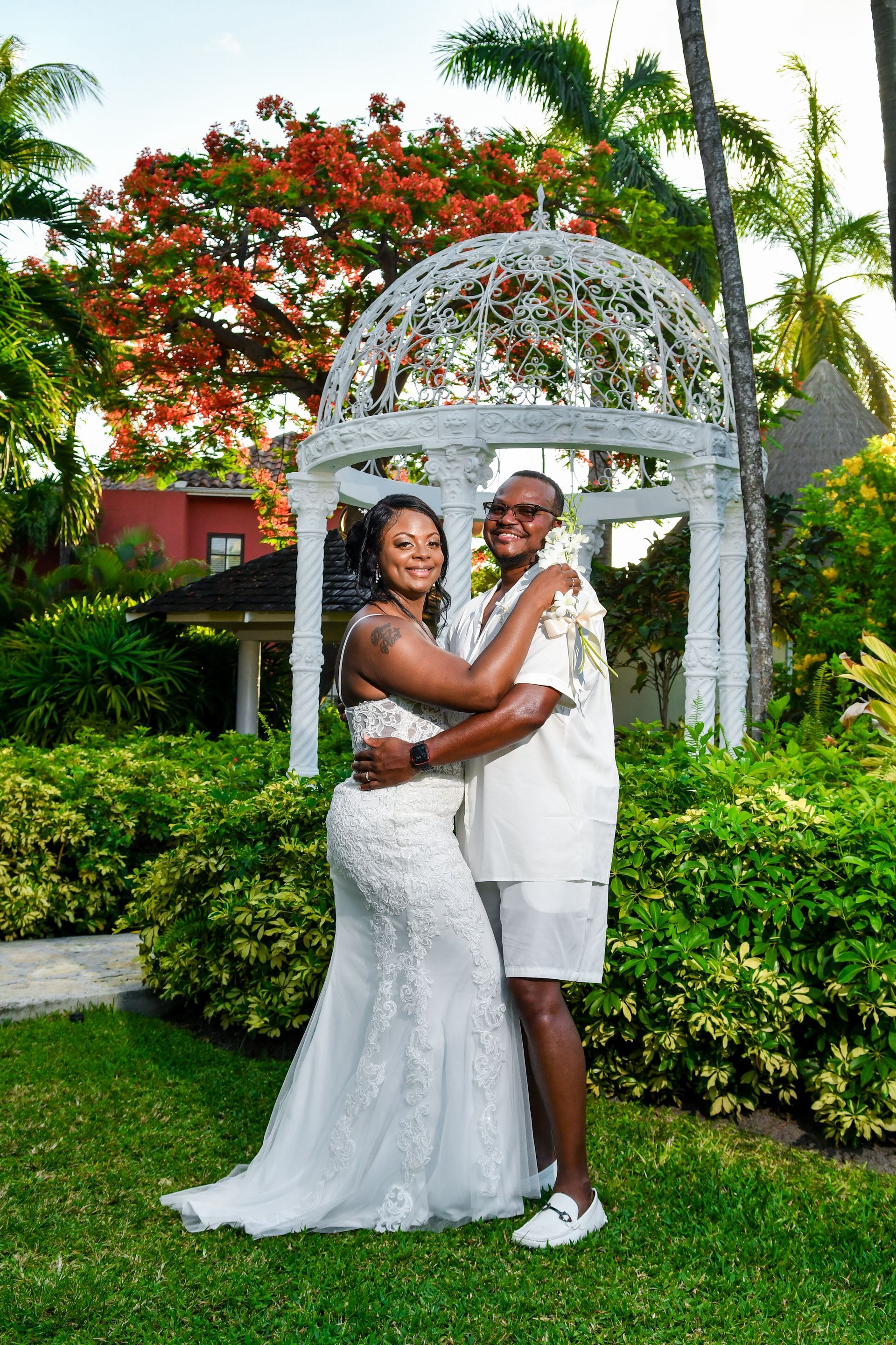 Two people embracing in white attire beneath a garden gazebo with lush greenery and flowers.