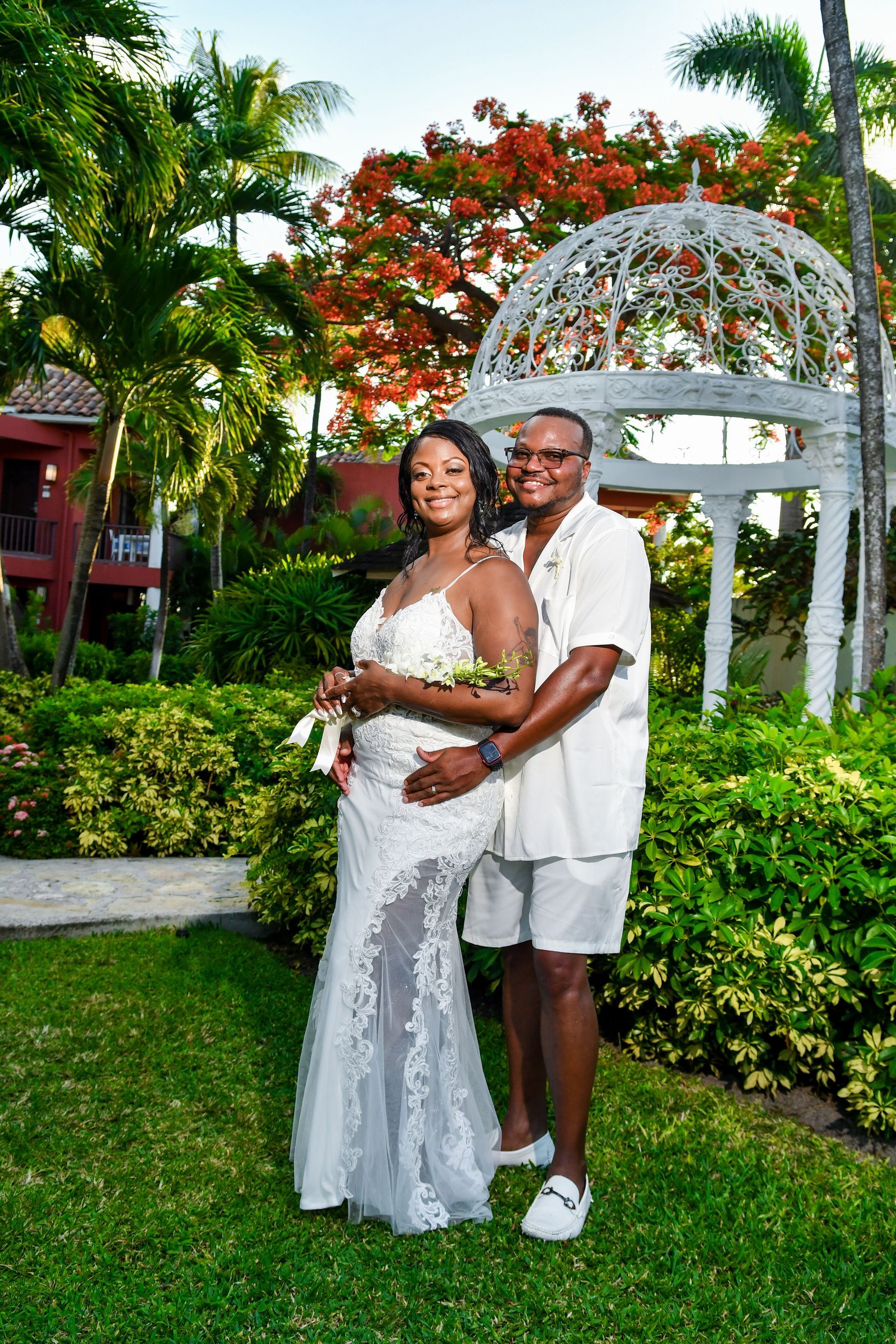 Couple in white outfits posing in a tropical garden under a white arbor