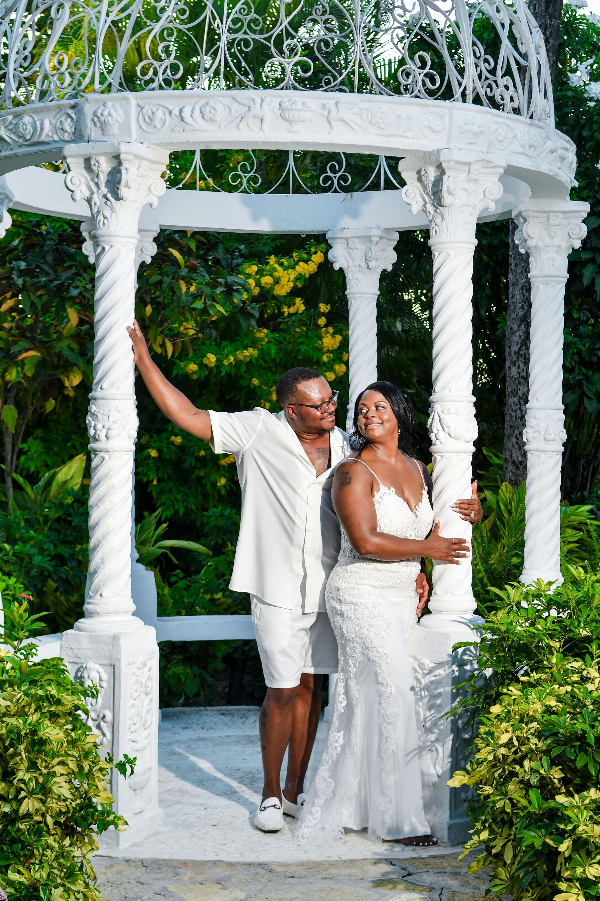 Couple in white standing under a white gazebo in a lush garden, posing together and smiling