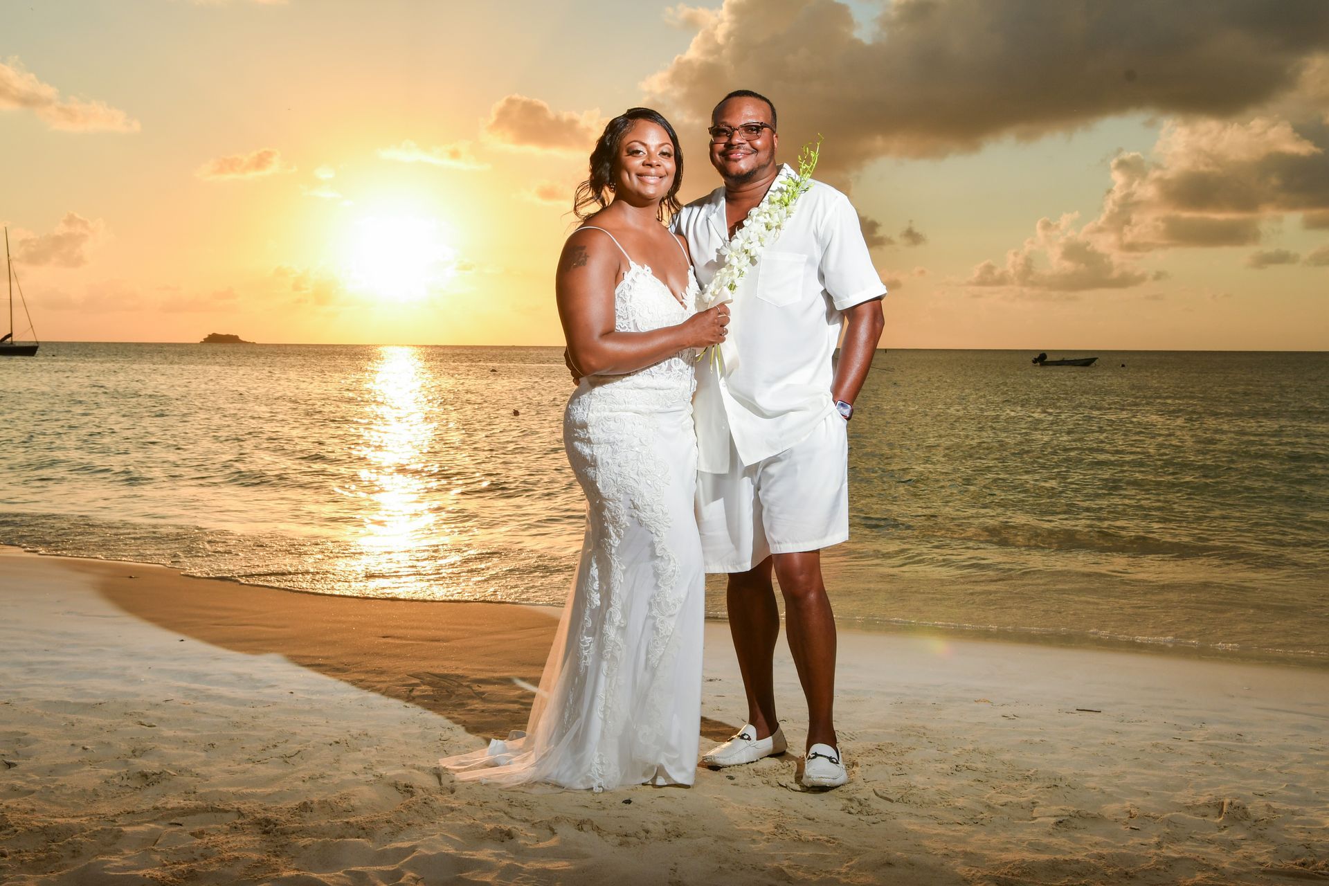 Couple in white wedding attire standing on a beach at sunset.