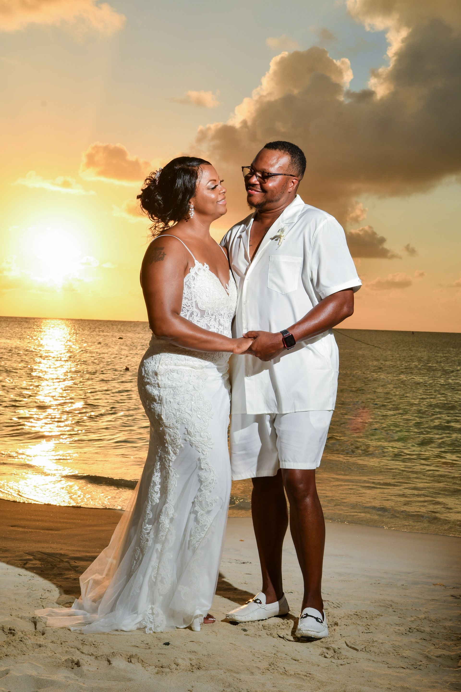 Couple in white standing on a beach at sunset, holding hands and smiling