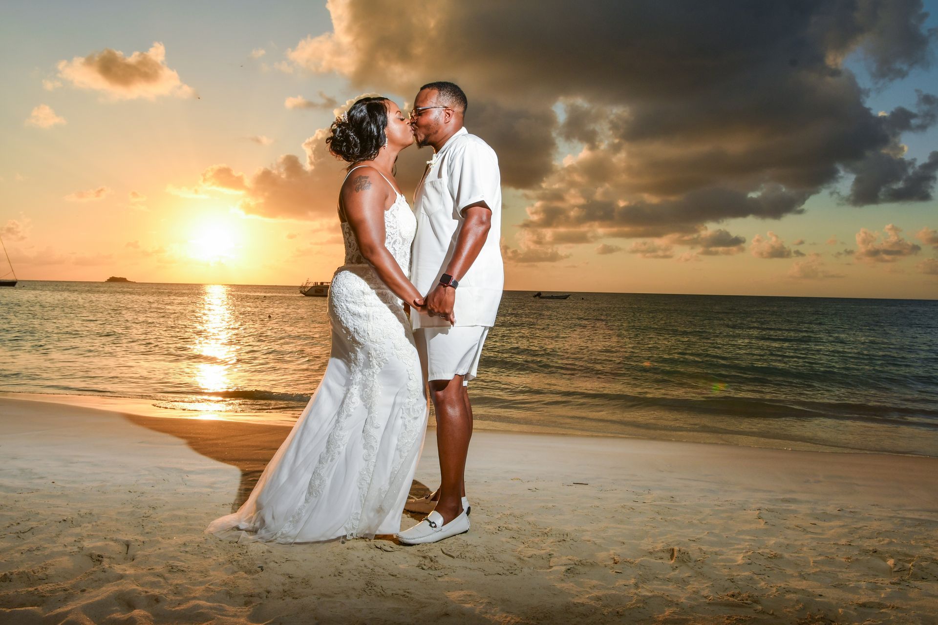Couple in white wedding attire kissing on a beach at sunset with waves and clouds in the background