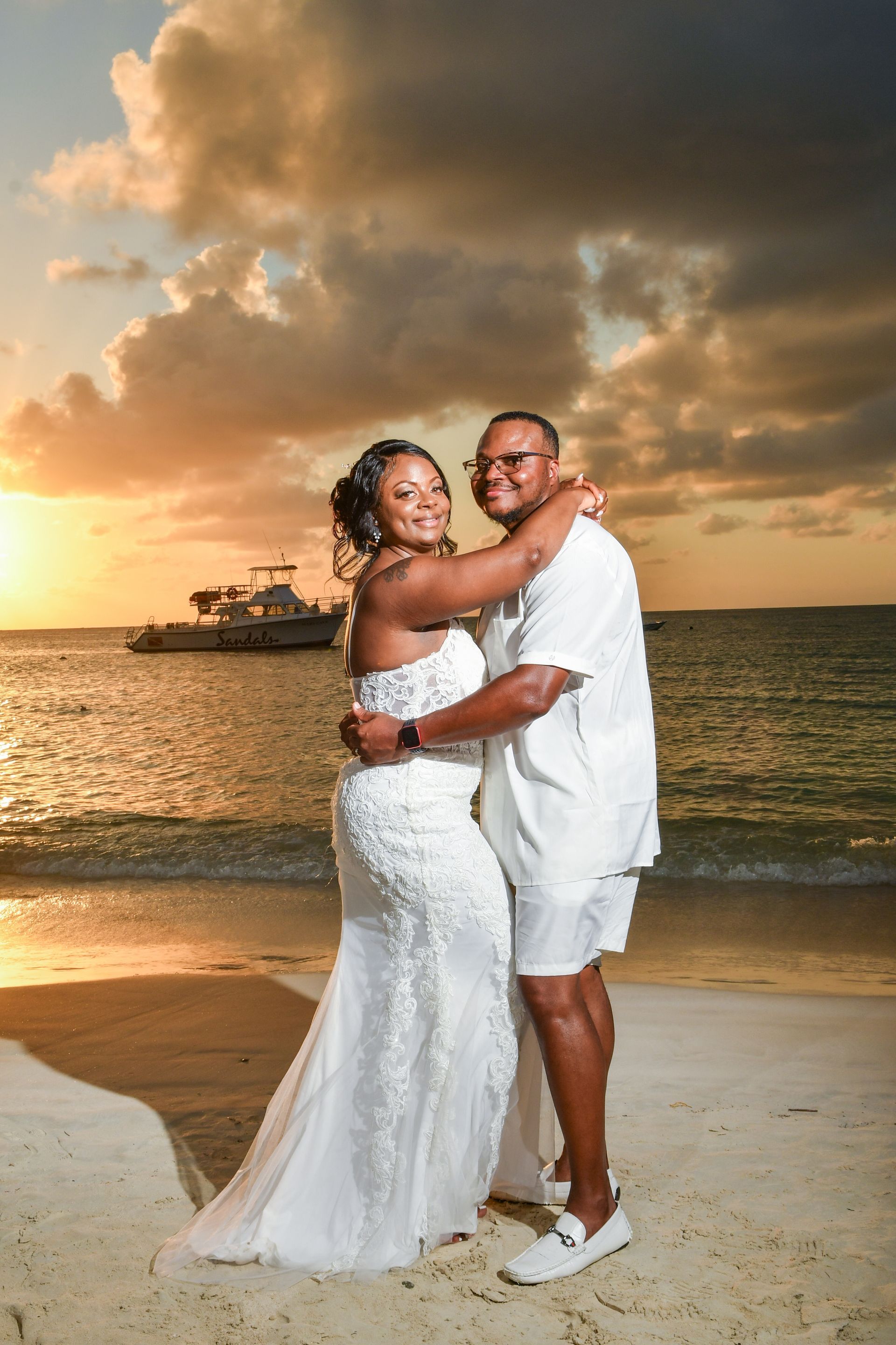 Couple embracing on a beach at sunset, with a boat in the background.