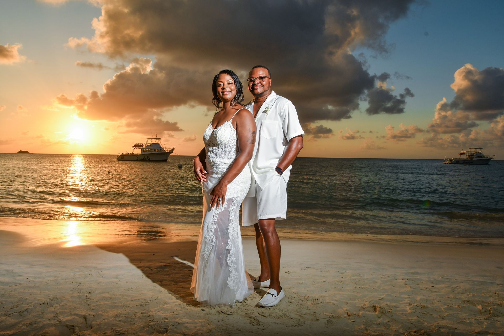Couple in white beachwear standing on a tropical beach at sunset, with boats and dramatic clouds behind them