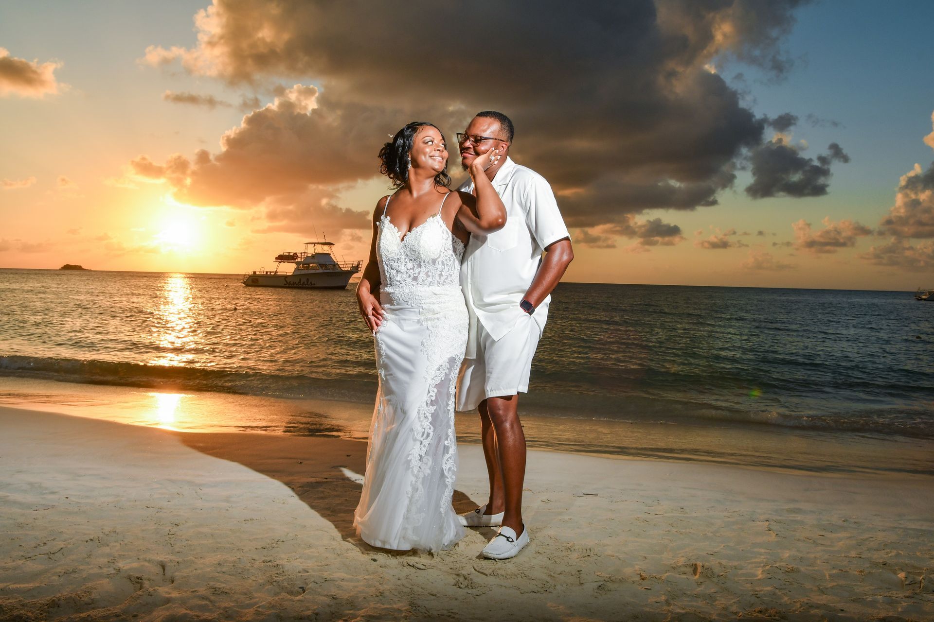 Couple in white outfits on a beach at sunset, standing by the water with dark clouds overhead