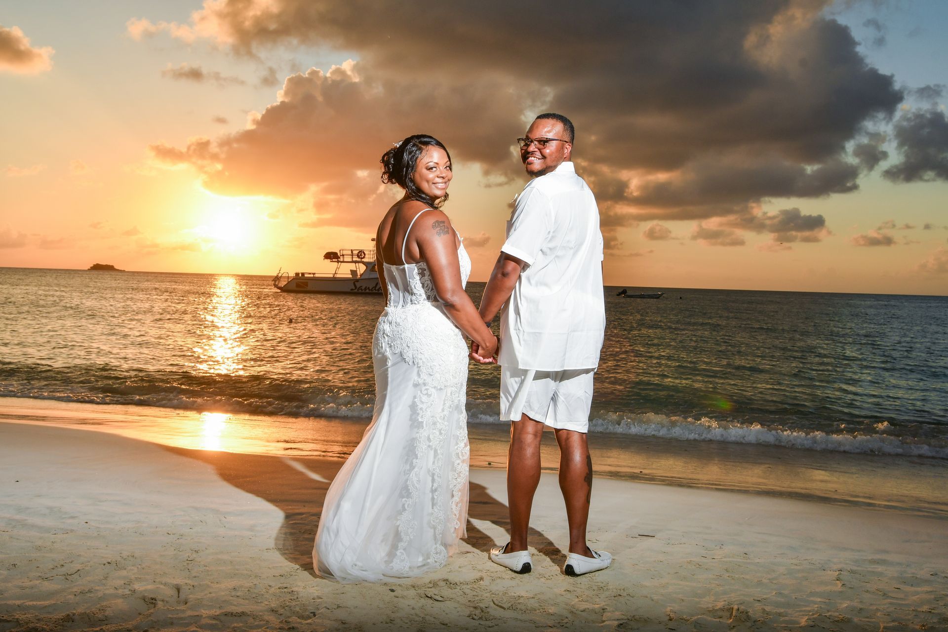 Couple in white outfits holding hands on a beach at sunset with dramatic clouds.