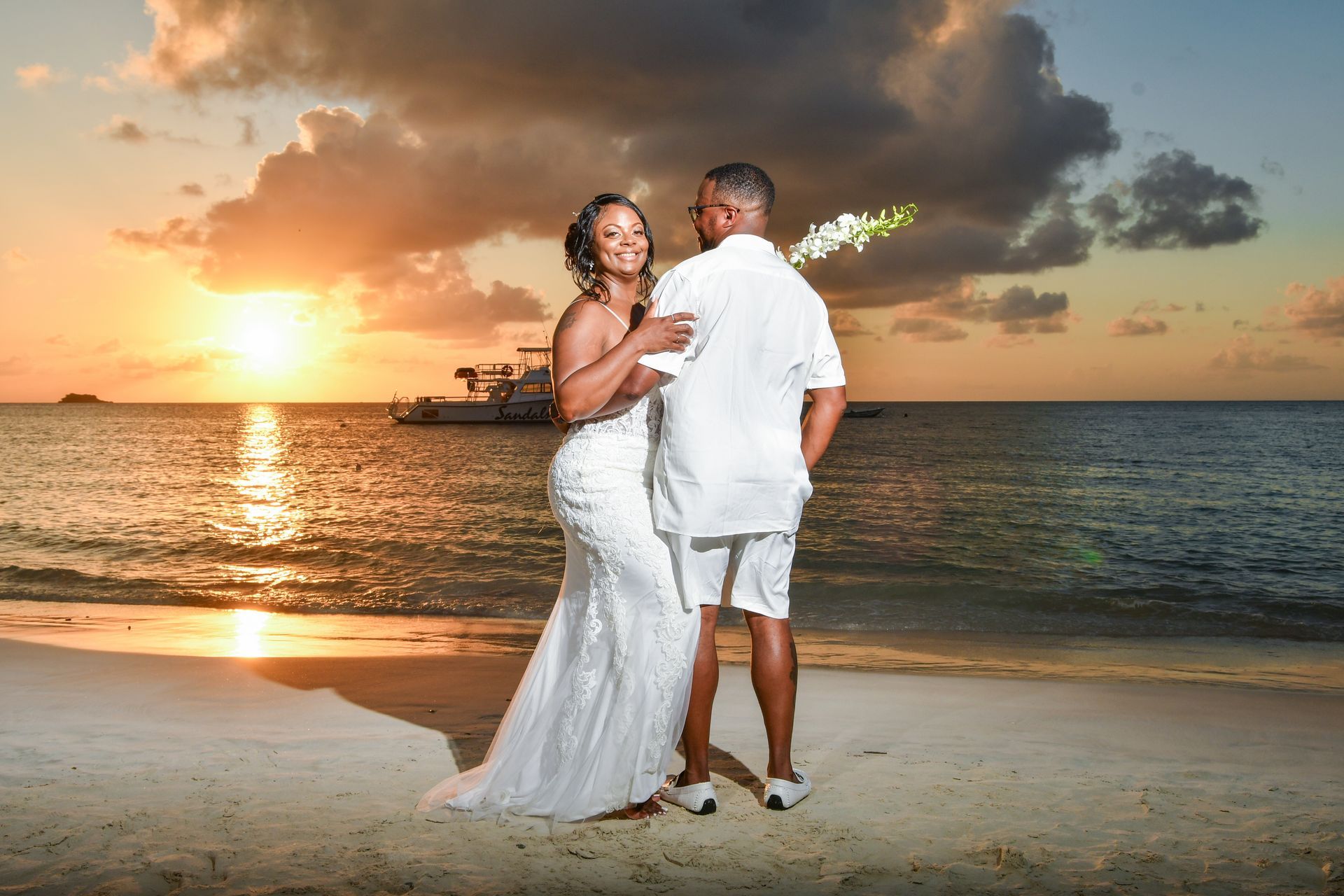 Couple in white outfits dancing on a beach at sunset, with ocean waves and warm sky behind them