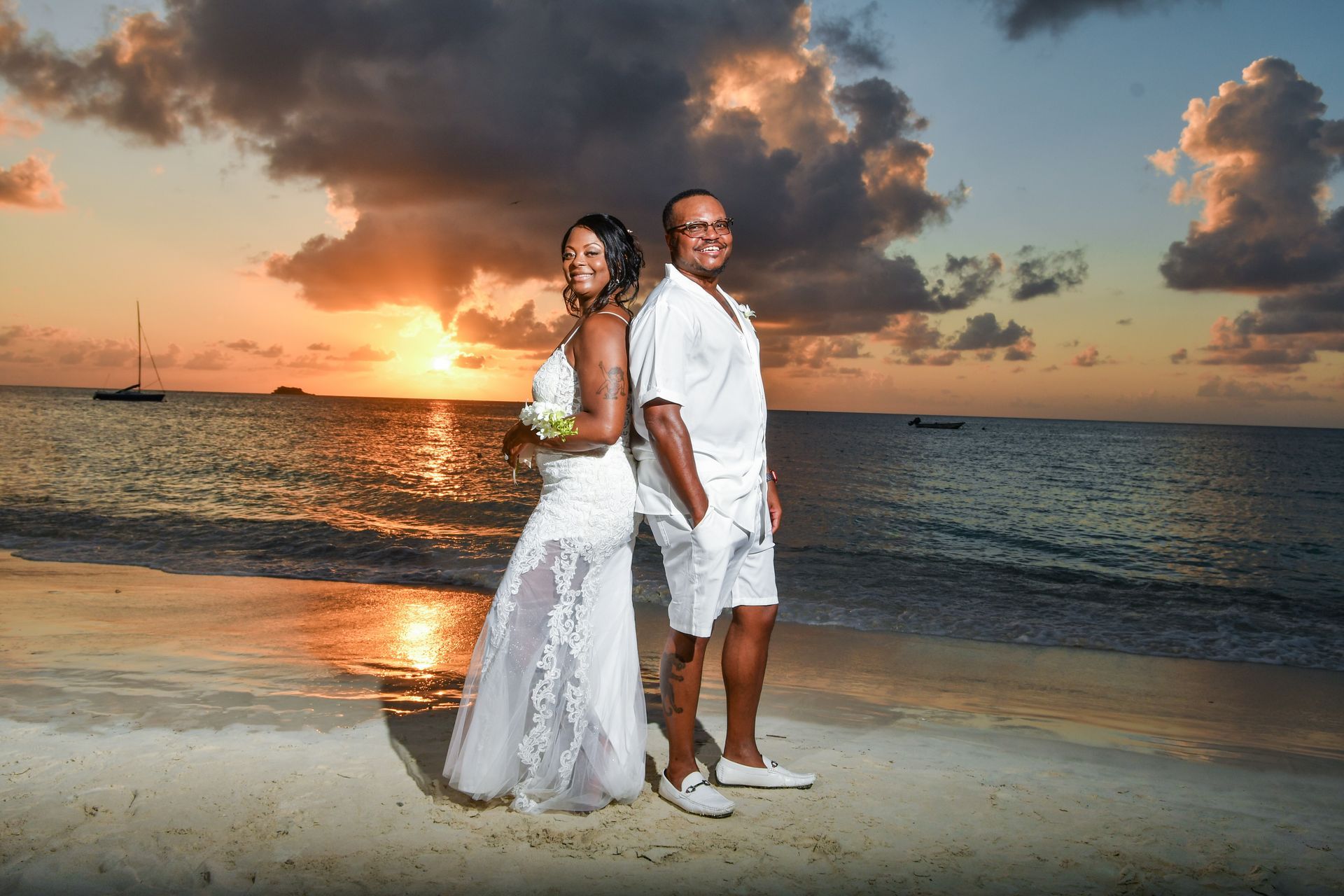 Couple in wedding attire standing on a beach at sunset, ocean and dramatic clouds behind them