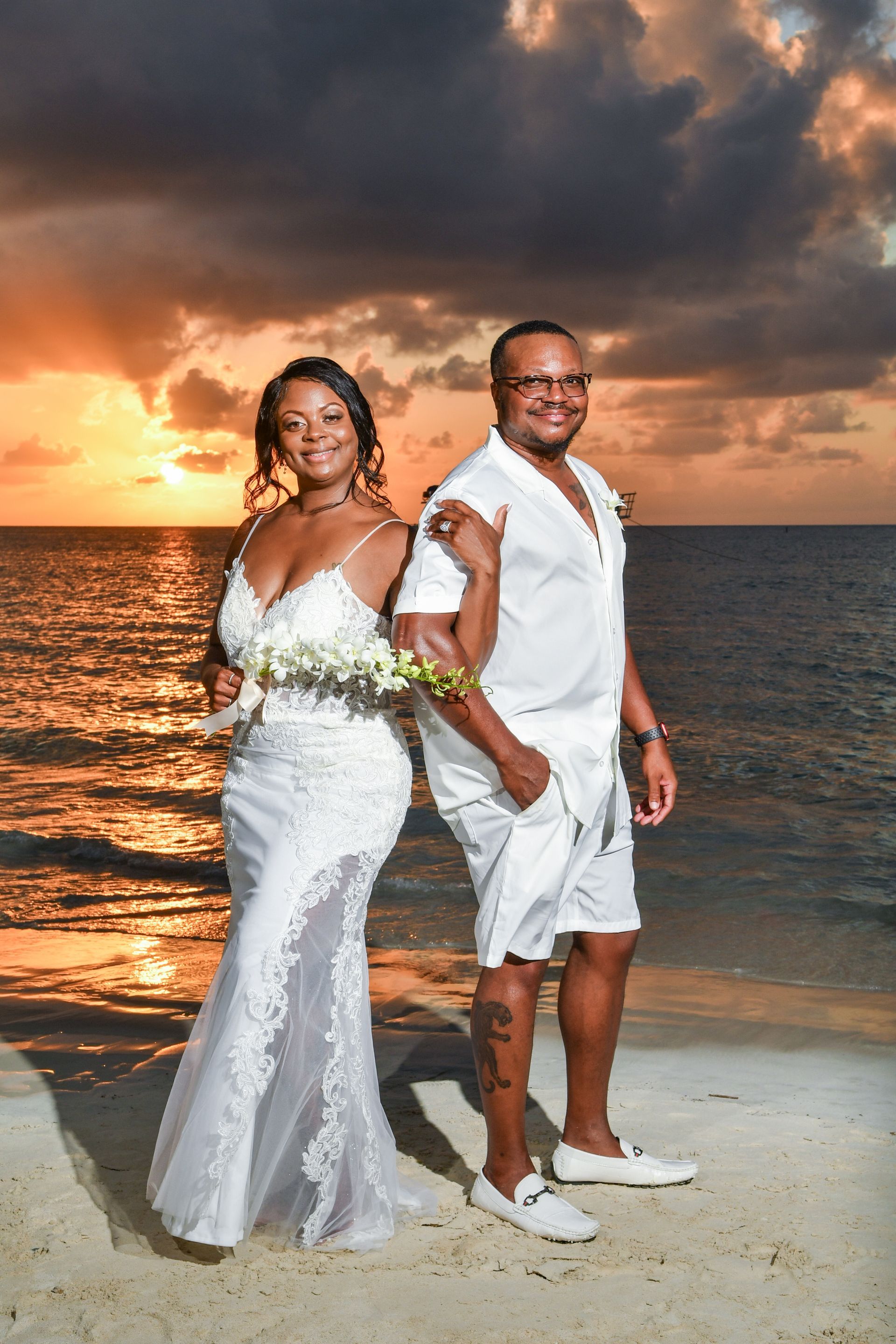 Couple in white beachwear holding hands on a beach at sunset, with dramatic clouds and ocean behind them.