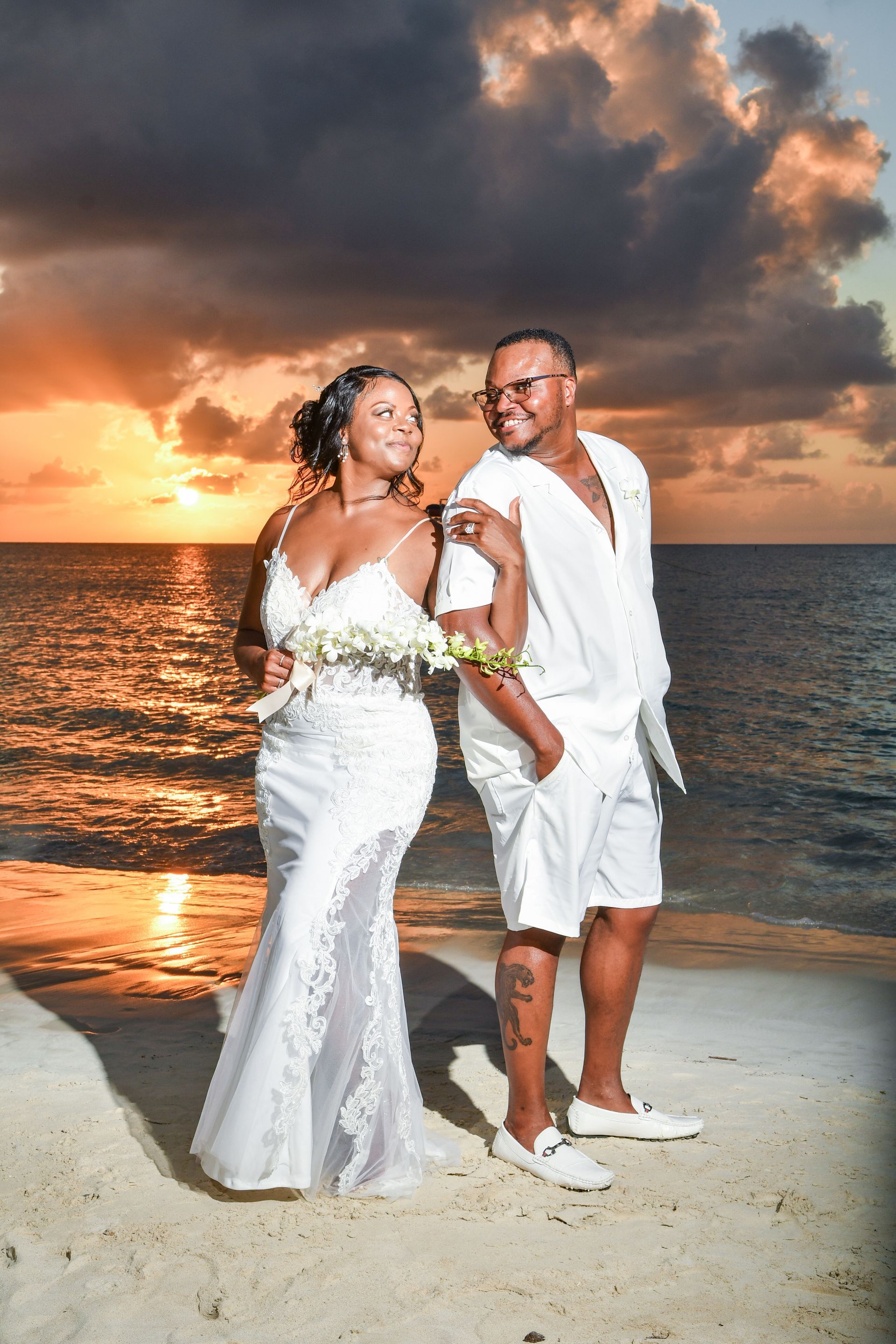 Couple in white outfits posing on a beach at sunset with golden water and dramatic clouds