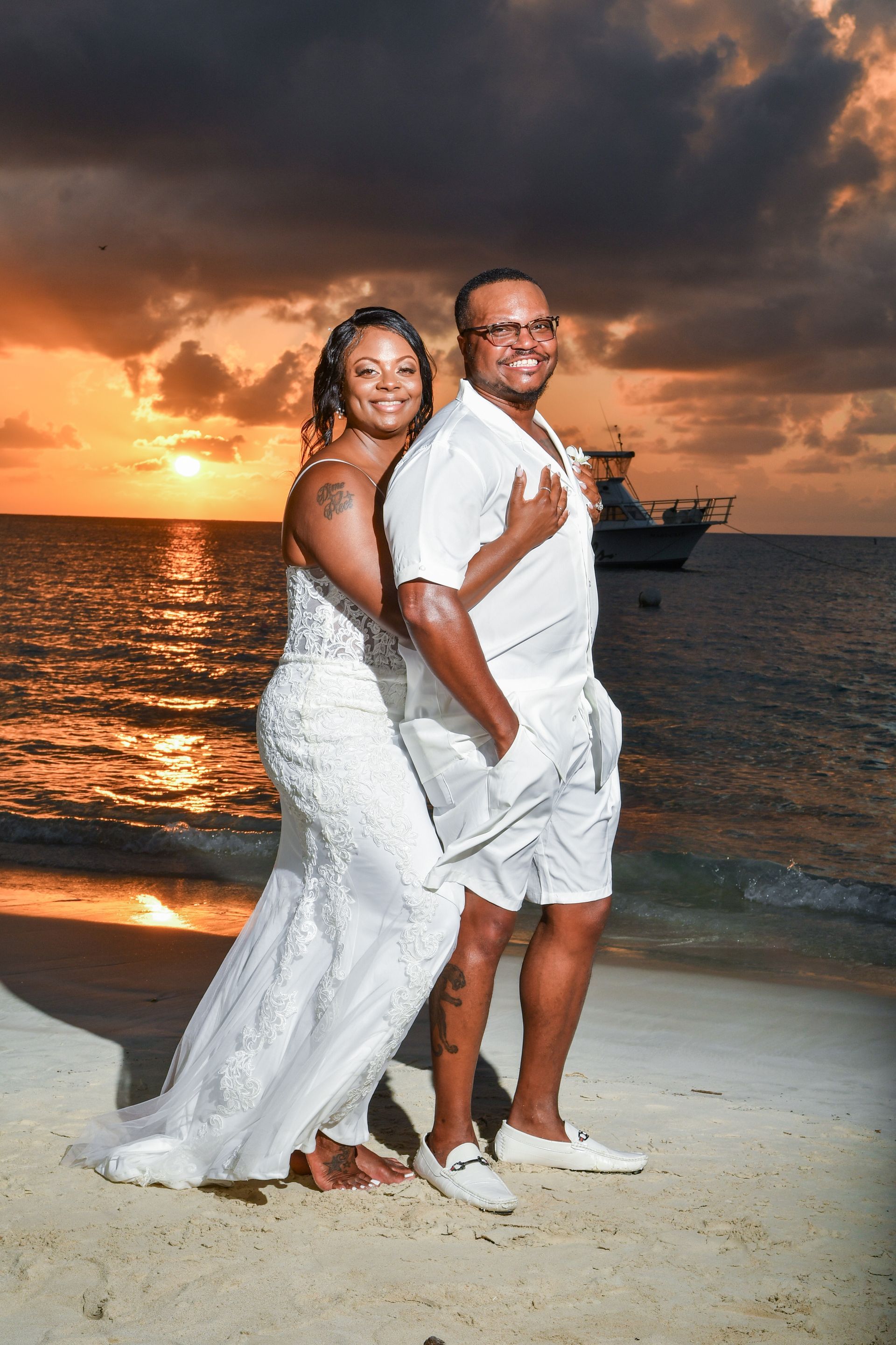 Couple in white outfits posing on a beach at sunset with orange sky and calm ocean behind them