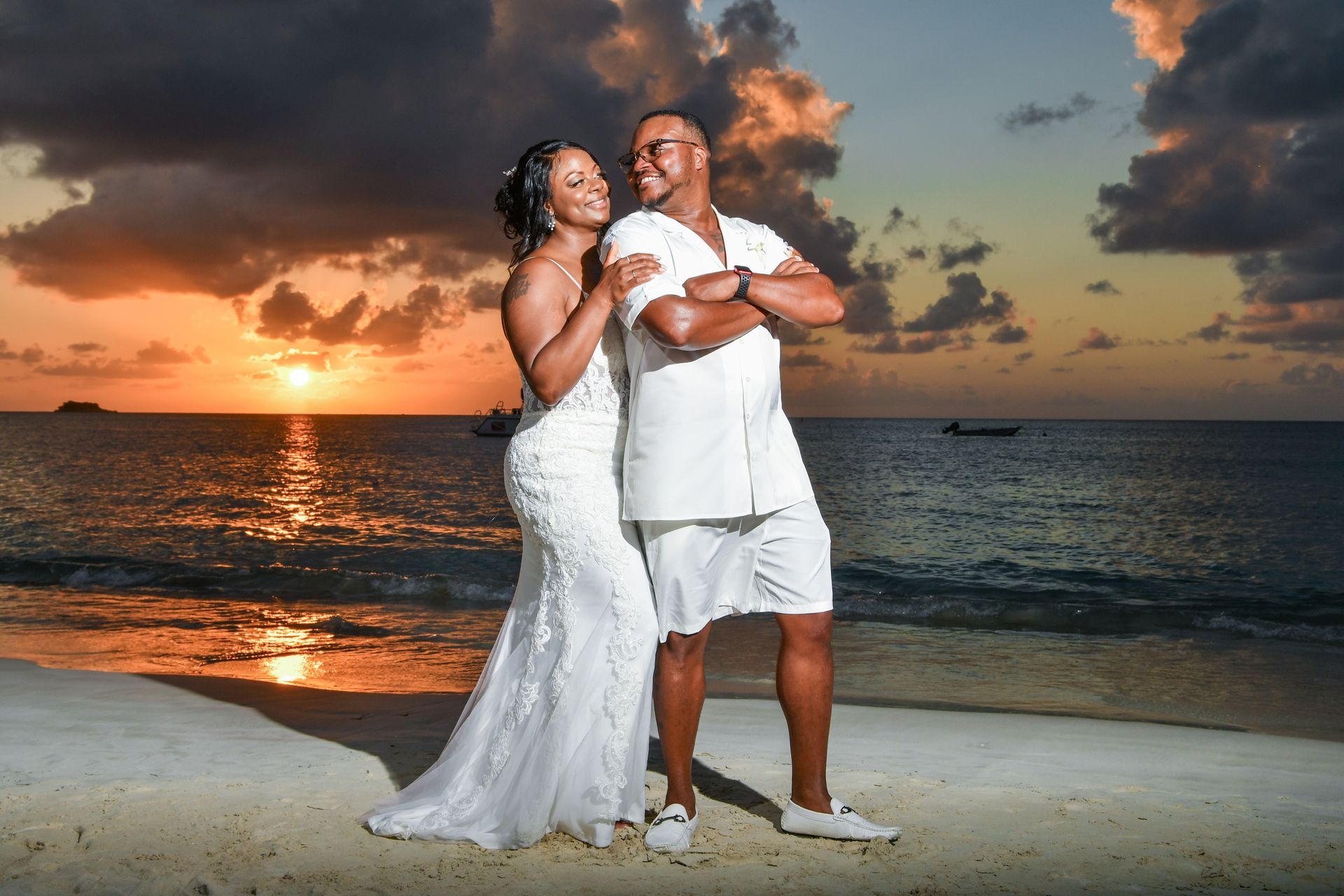 Couple in white standing on a beach at sunset, with orange clouds and calm ocean waves.