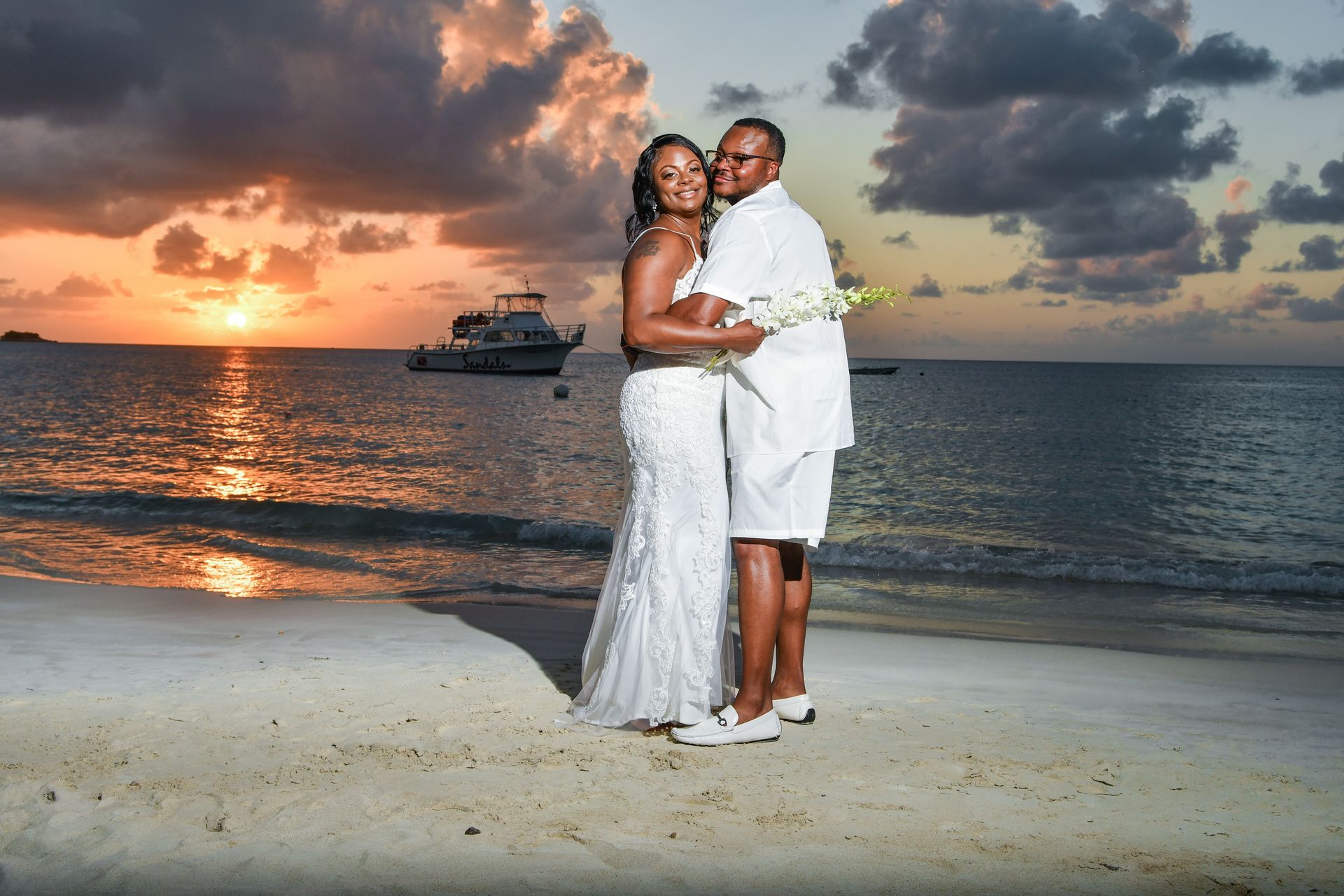 Couple in white outfits posing on a beach at sunset with a boat on the horizon.