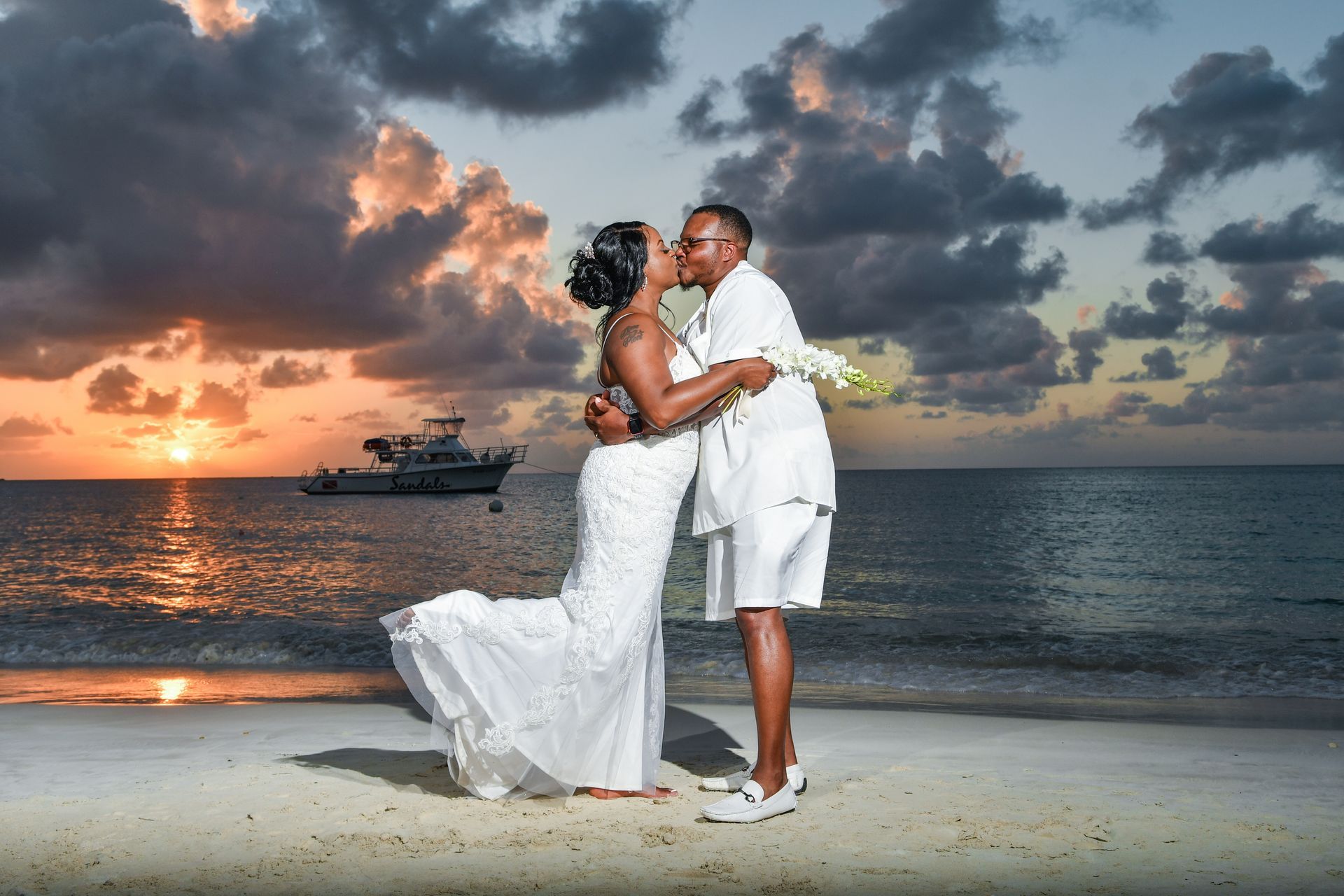 Couple in white embrace on a beach at sunset with ocean and boat in the background