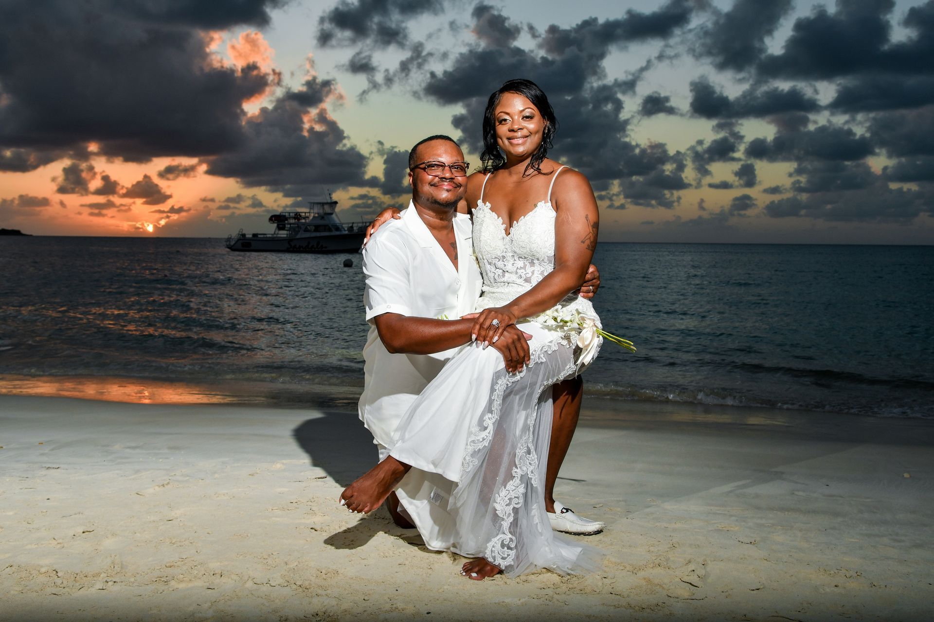 Couple in white formalwear sitting on a beach at sunset with the ocean and dramatic clouds behind them