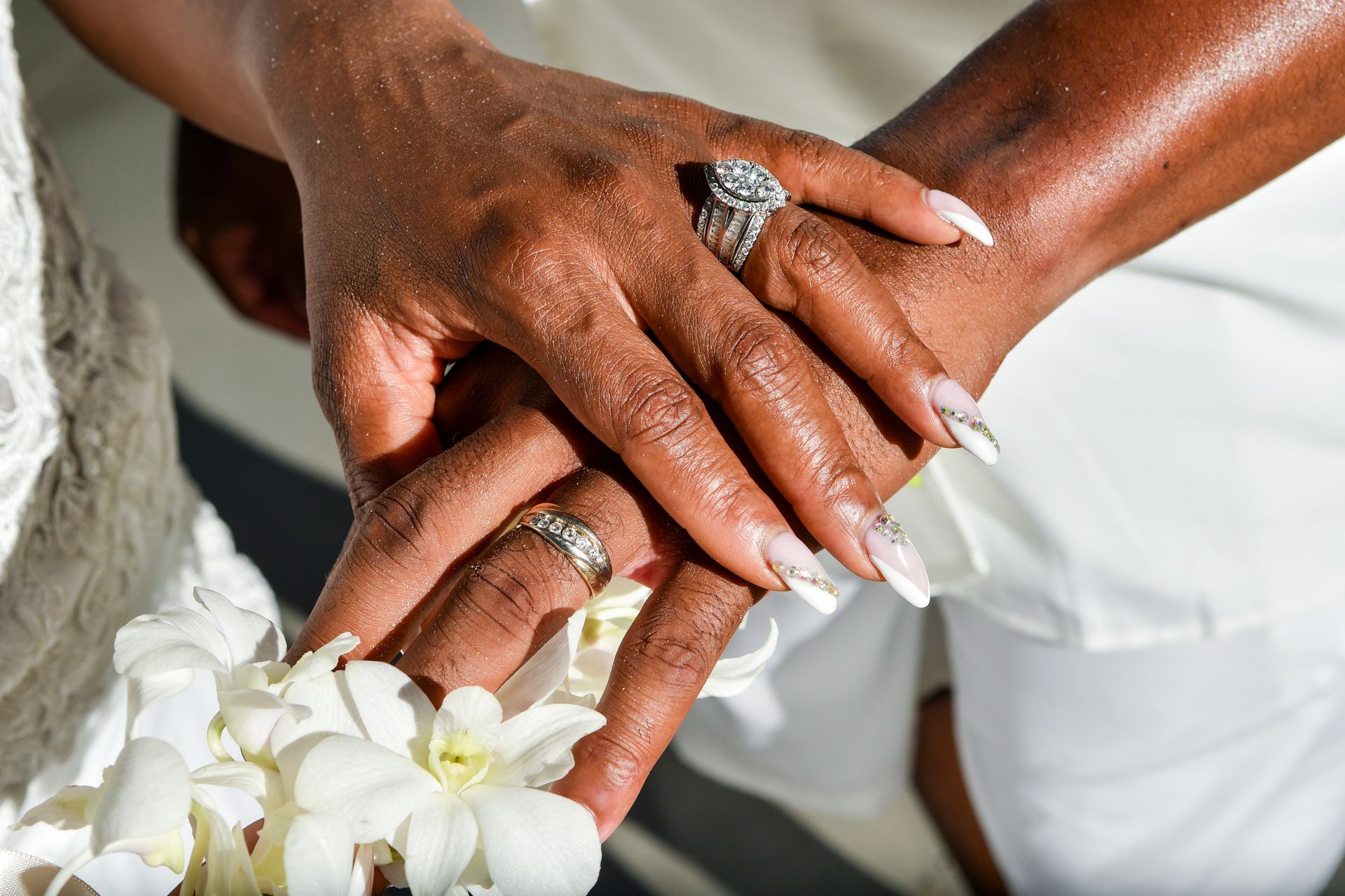 Hands with wedding rings resting on white flowers, with a white dress in the background