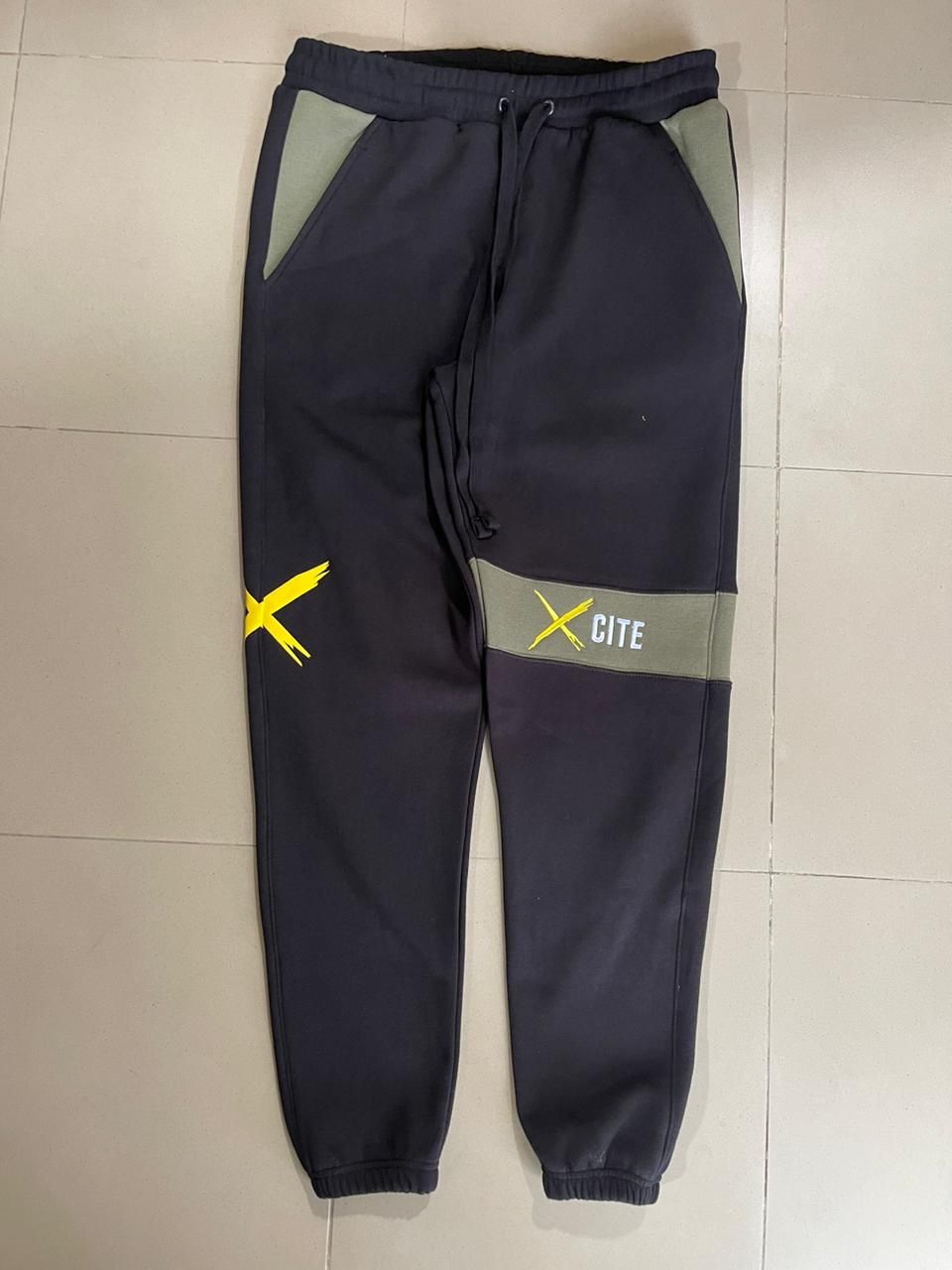 A pair of black sweatpants with a yellow x on the side are sitting on a tiled floor.