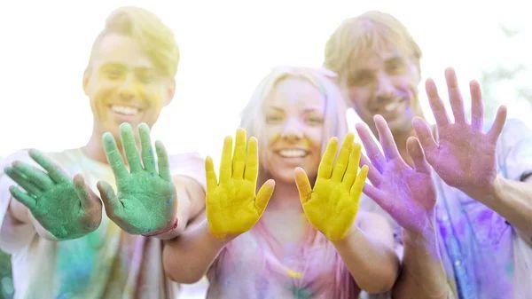 Three people smiling, holding up colorful hands. Yellow, green, and purple powder covers them.
