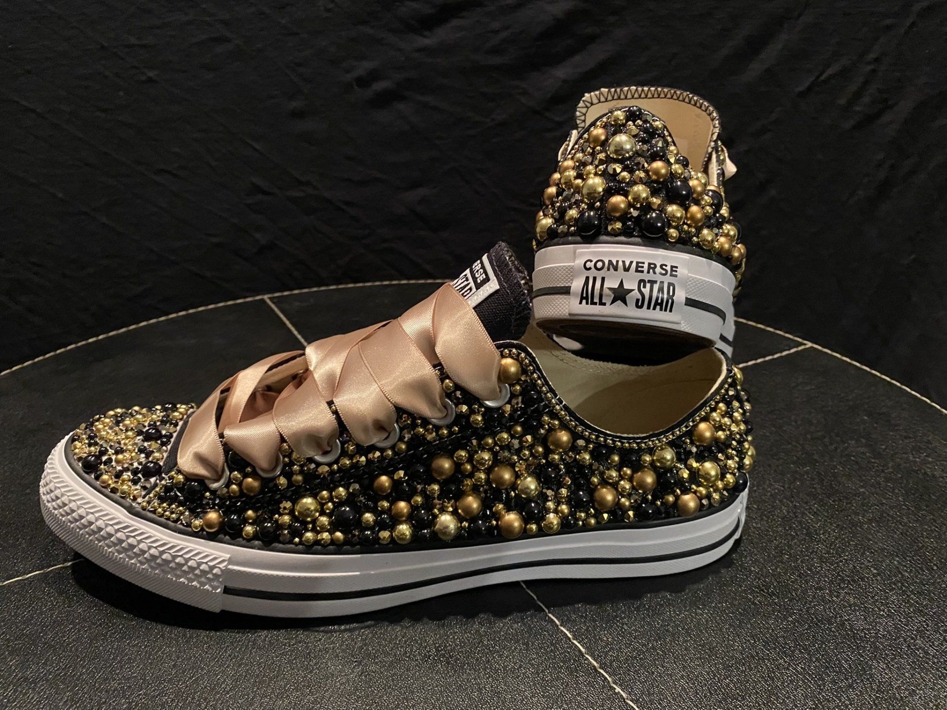 Black Converse sneaker bedazzled with gold beads, gold ribbon laces, and a matching ankle cuff on a black background.