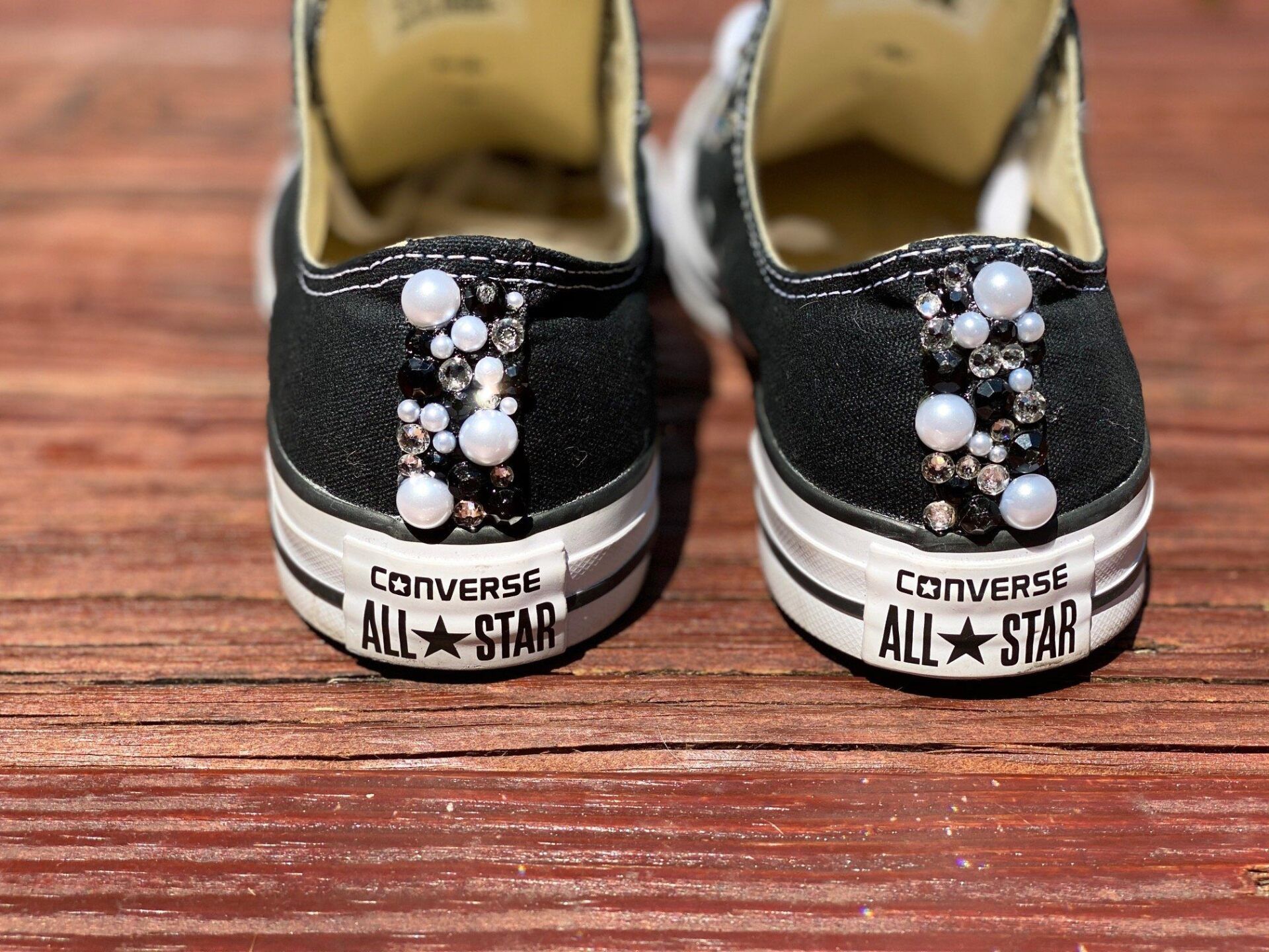 Black Converse sneakers with pearl and crystal embellishments on the heel, sitting on a wooden surface.