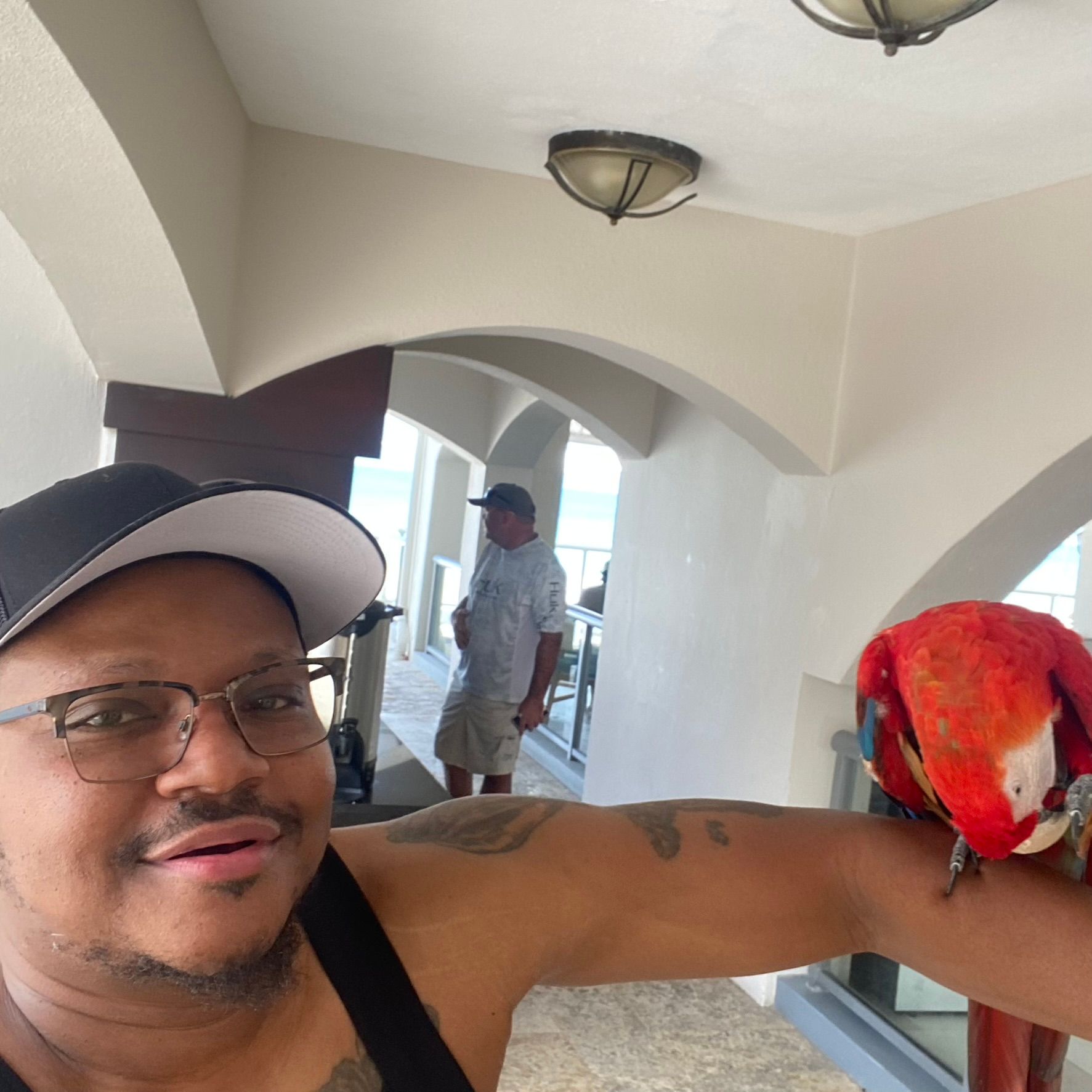 Man with glasses and cap holding a red parrot on his arm in a bright indoor hallway