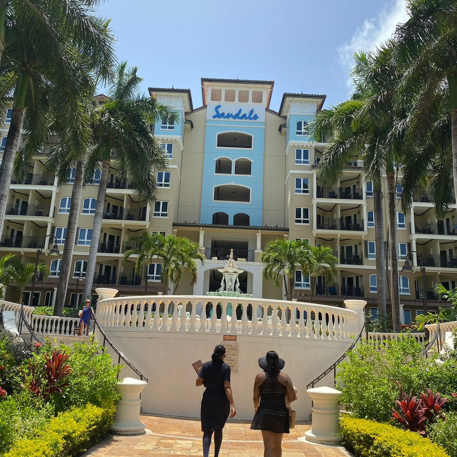 Two people standing before a pastel resort building with palm trees and blue sky
