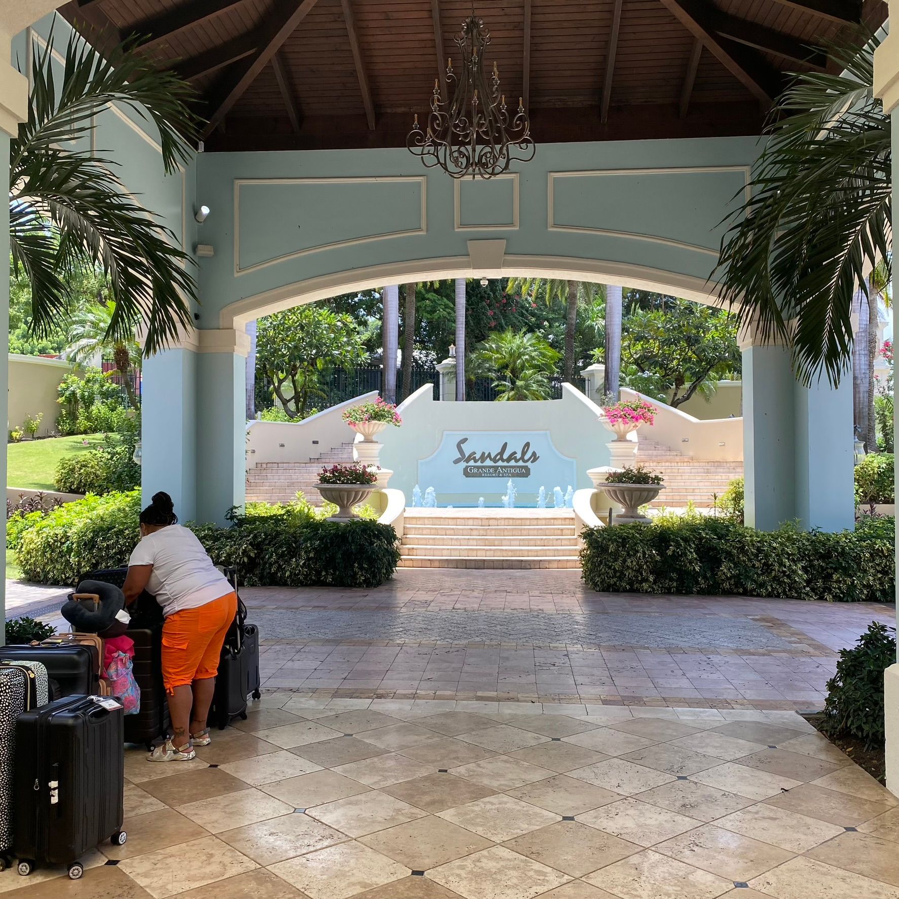 Resort entrance with covered walkway, luggage cart, and white sign framed by tropical plants