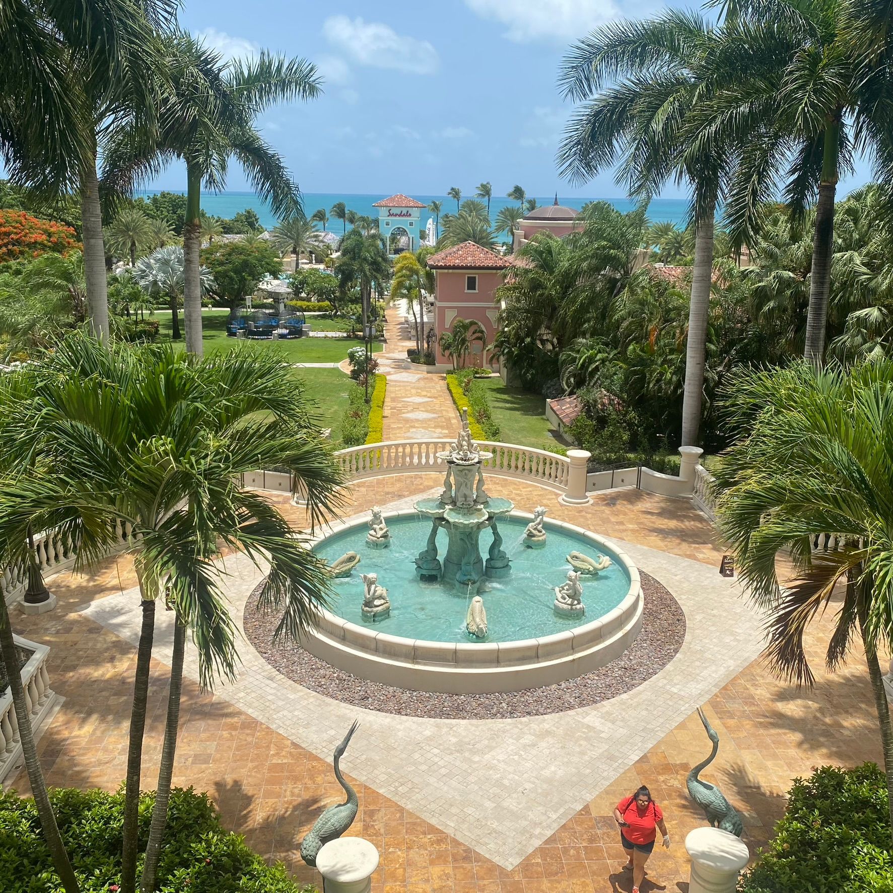 Garden courtyard with a blue fountain, palm trees, and a walkway leading to a small building under a bright sky