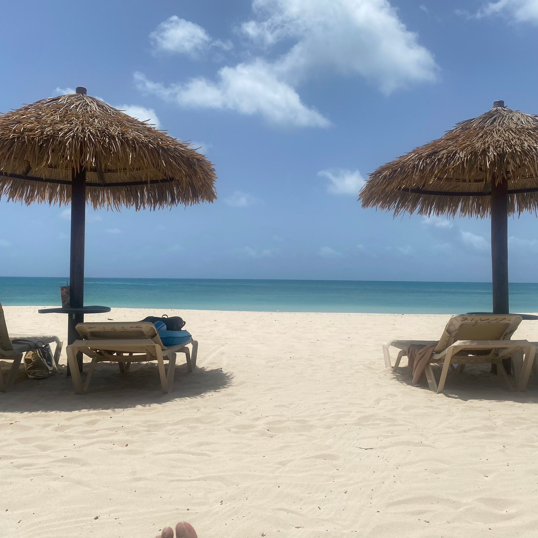 Beach scene with two straw umbrellas, lounge chairs, white sand, and turquoise ocean under a blue sky.