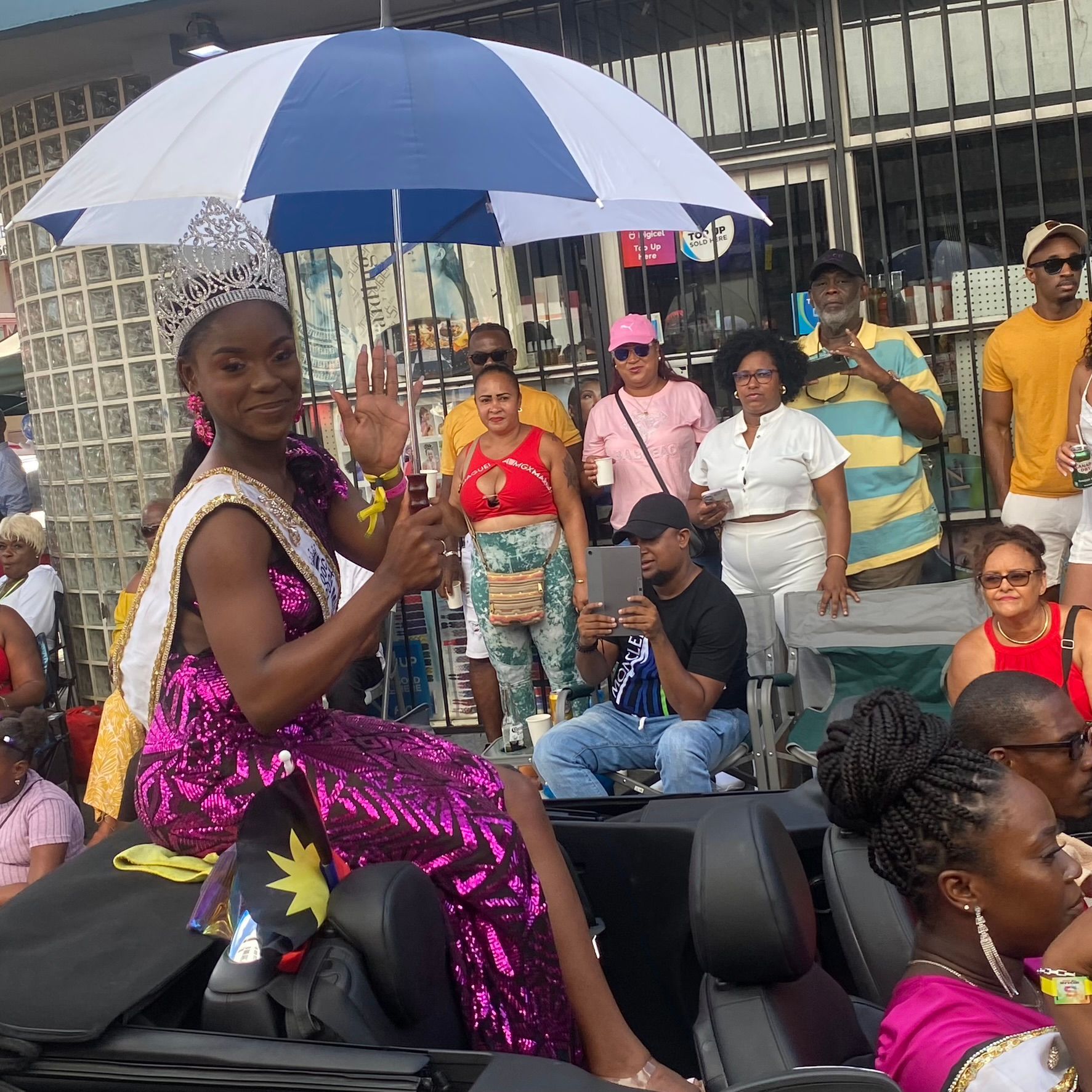 Person riding on another person’s shoulders under a blue-and-white umbrella at an outdoor crowd event