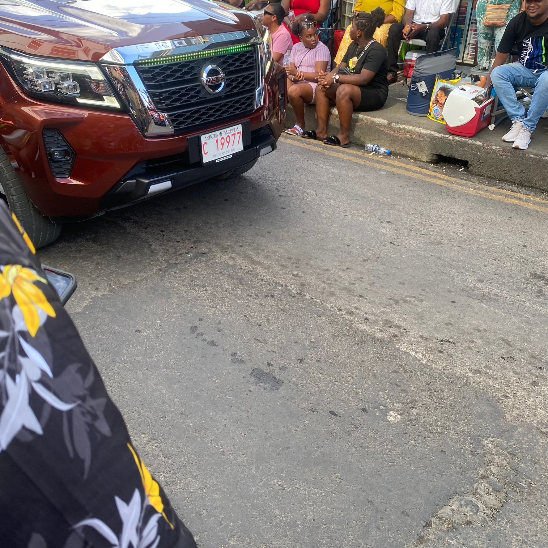Maroon SUV stopped on a street beside seated pedestrians near a market stall.