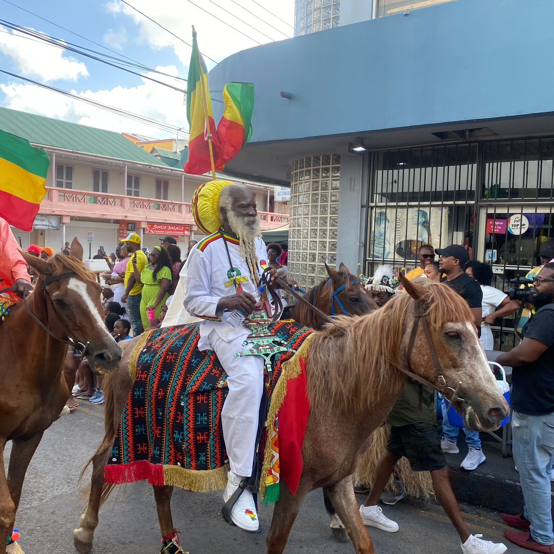 Person in white rides a brown horse in a colorful street parade, with flags and onlookers nearby.