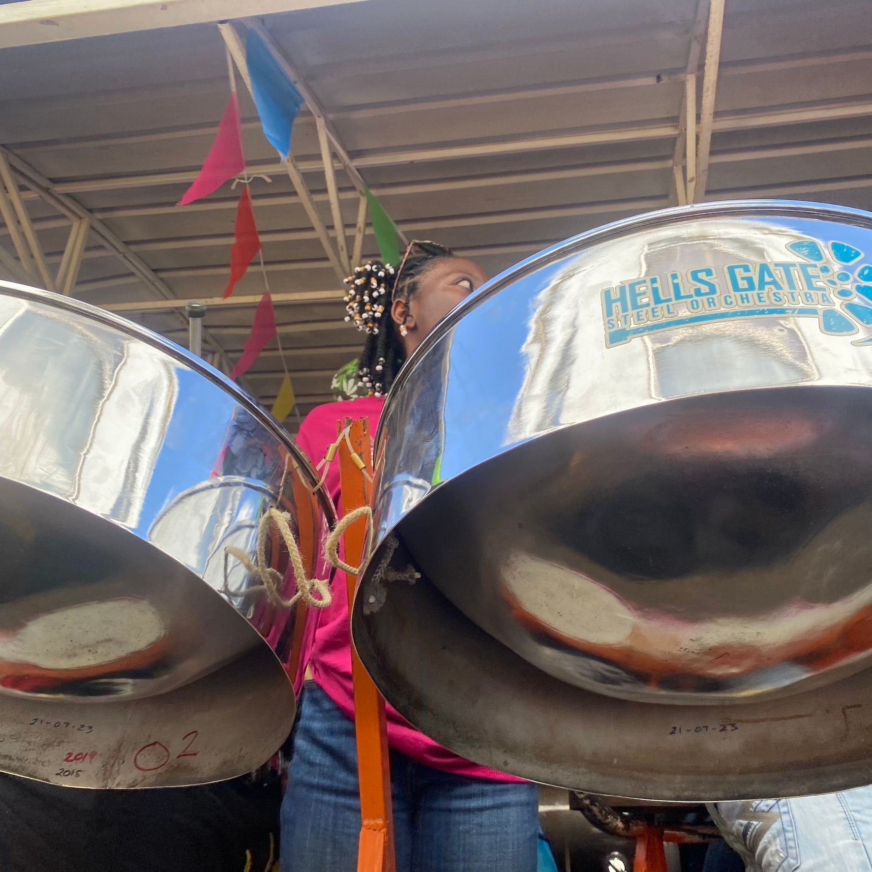 Person playing shiny steel drums outdoors under colorful flags