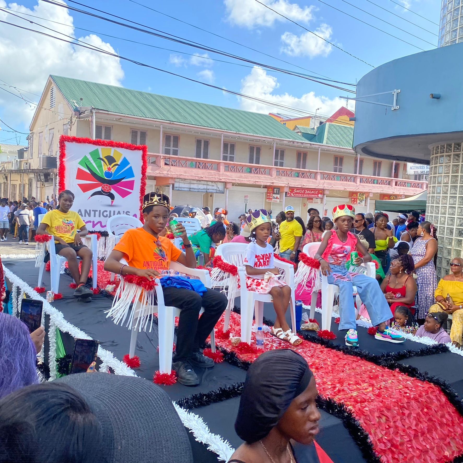 People seated on a decorated outdoor float during a colorful parade, with flags and spectators nearby.