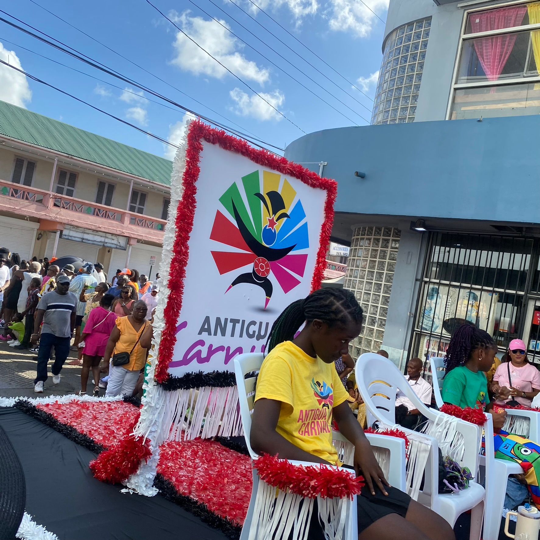 Street parade float with “Antigua” sign, red-white decorations, and seated people in a sunny town setting