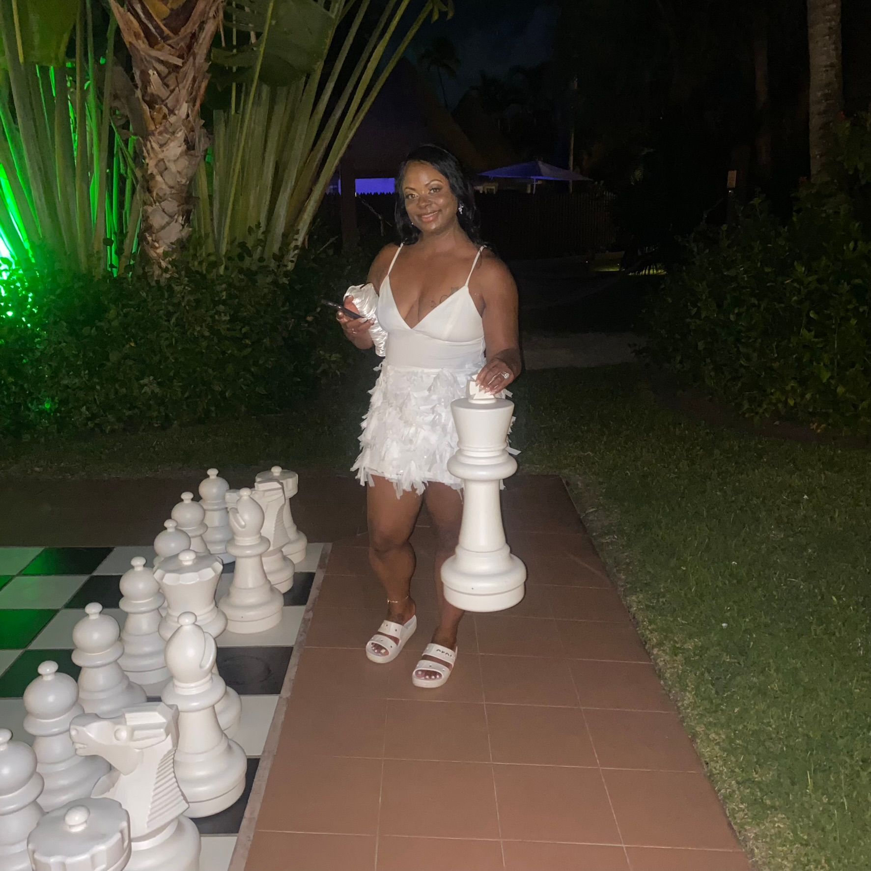 Woman in a white dress posing beside oversized chess pieces on a garden walkway at night