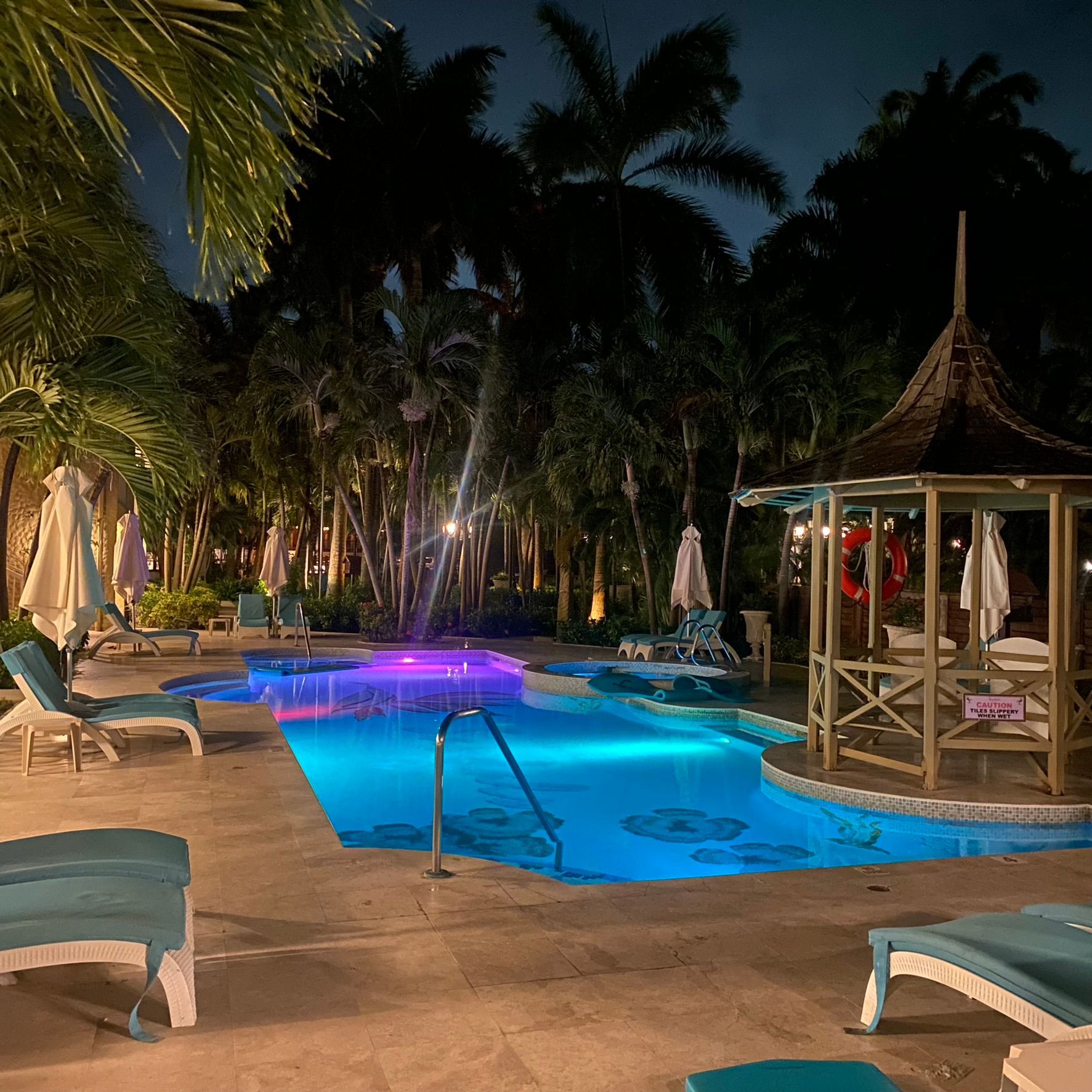 Tropical resort pool at night with blue lighting, lounge chairs, palm trees, and a gazebo