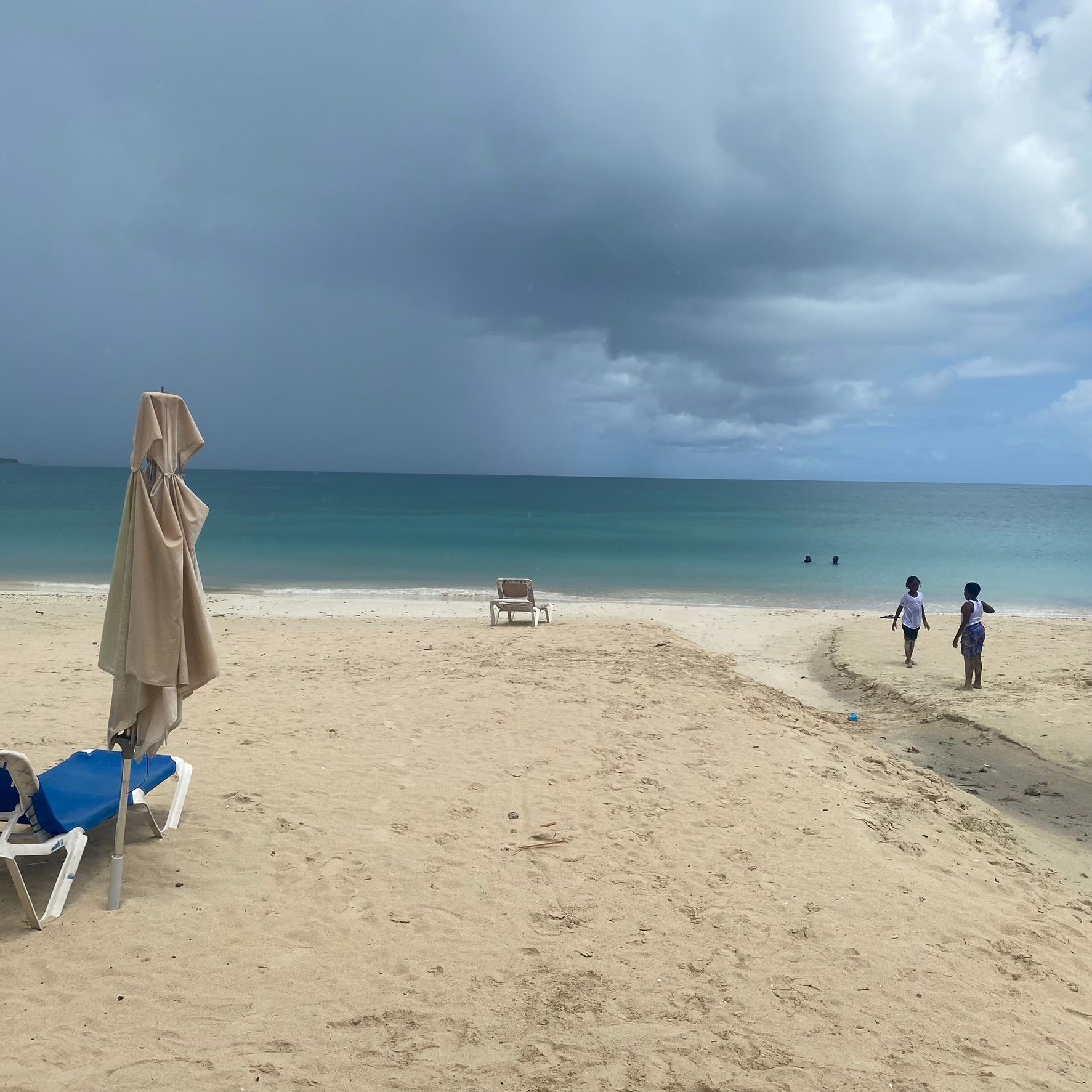 Sandy beach with turquoise water, storm clouds, a lone chair, and two people walking near the shoreline.