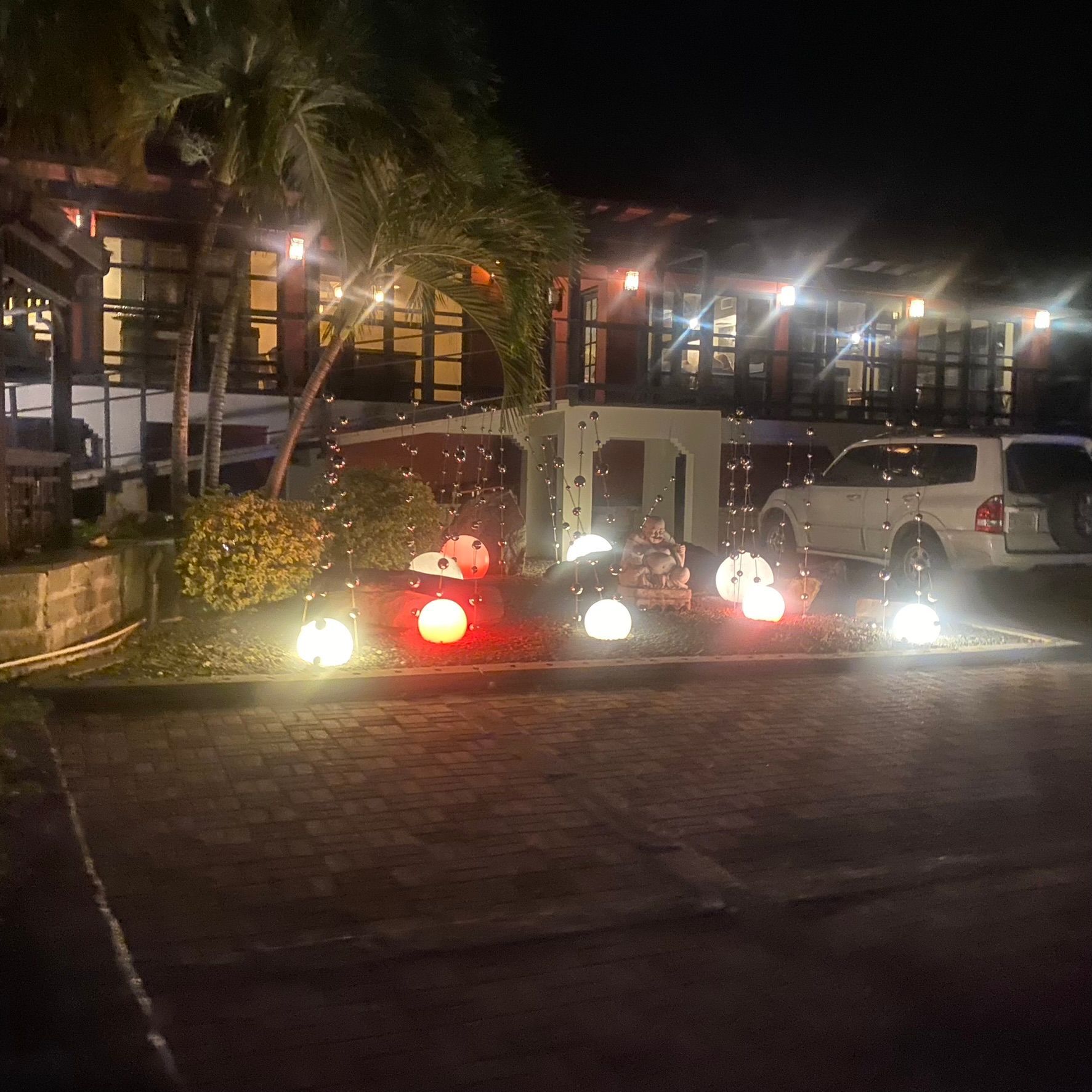 Nighttime street scene with a lit building, parked cars, and a red car with bright headlights in front.