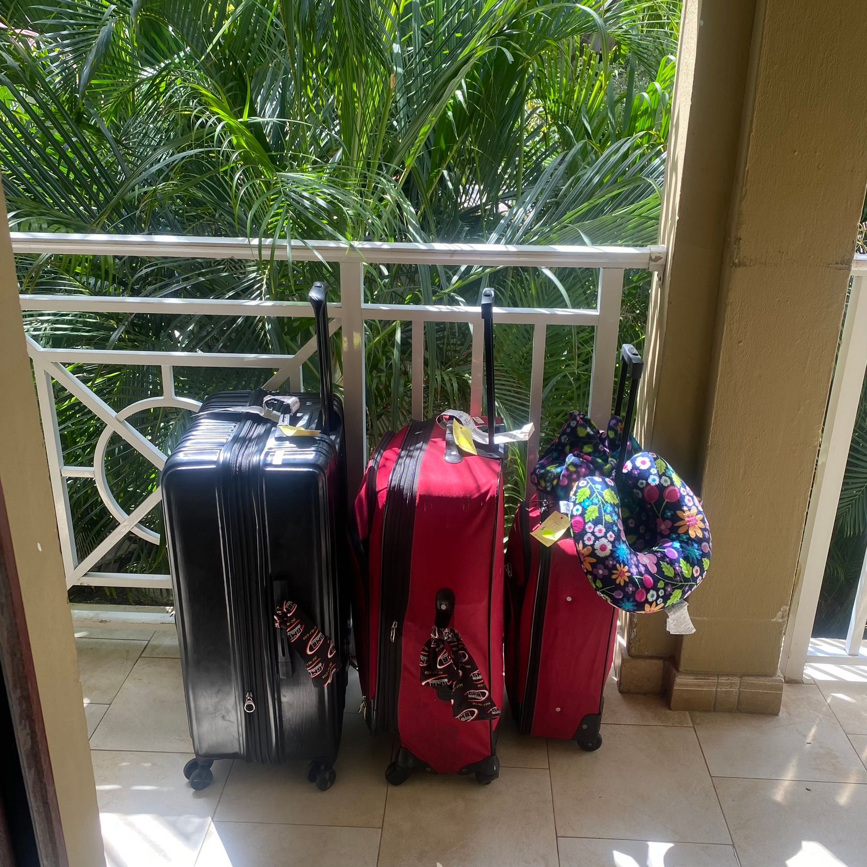 Three upright suitcases—black, red, and red—with a floral hat on a balcony beside a white railing.