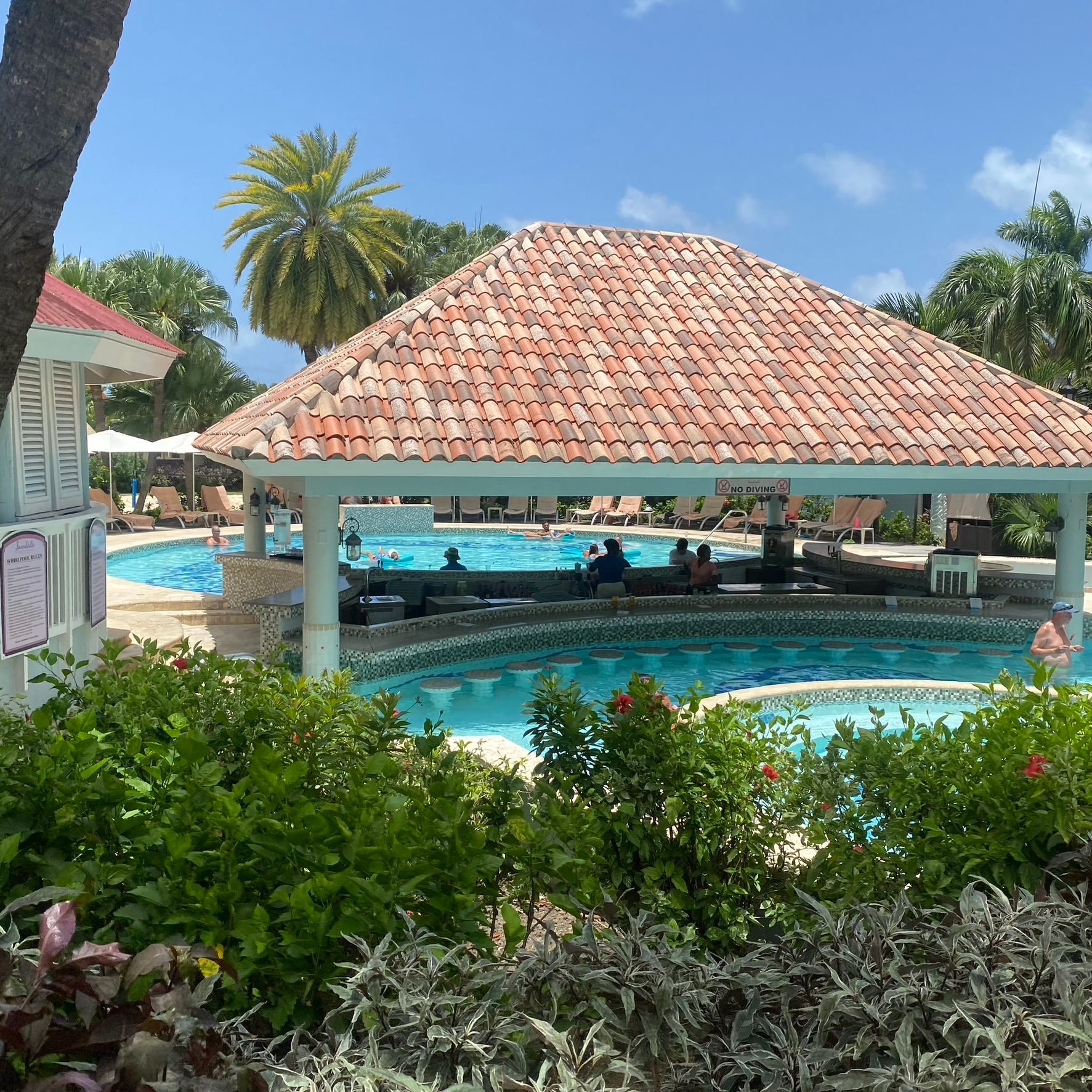 Tropical resort pool with a red-tiled pavilion, palm trees, and bright blue water under a clear sky