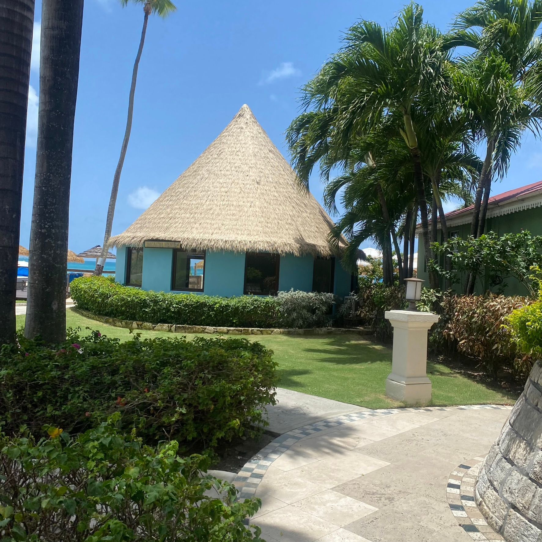 Tropical resort bungalow with thatched roof, palm trees, and a sunlit garden path under a clear blue sky
