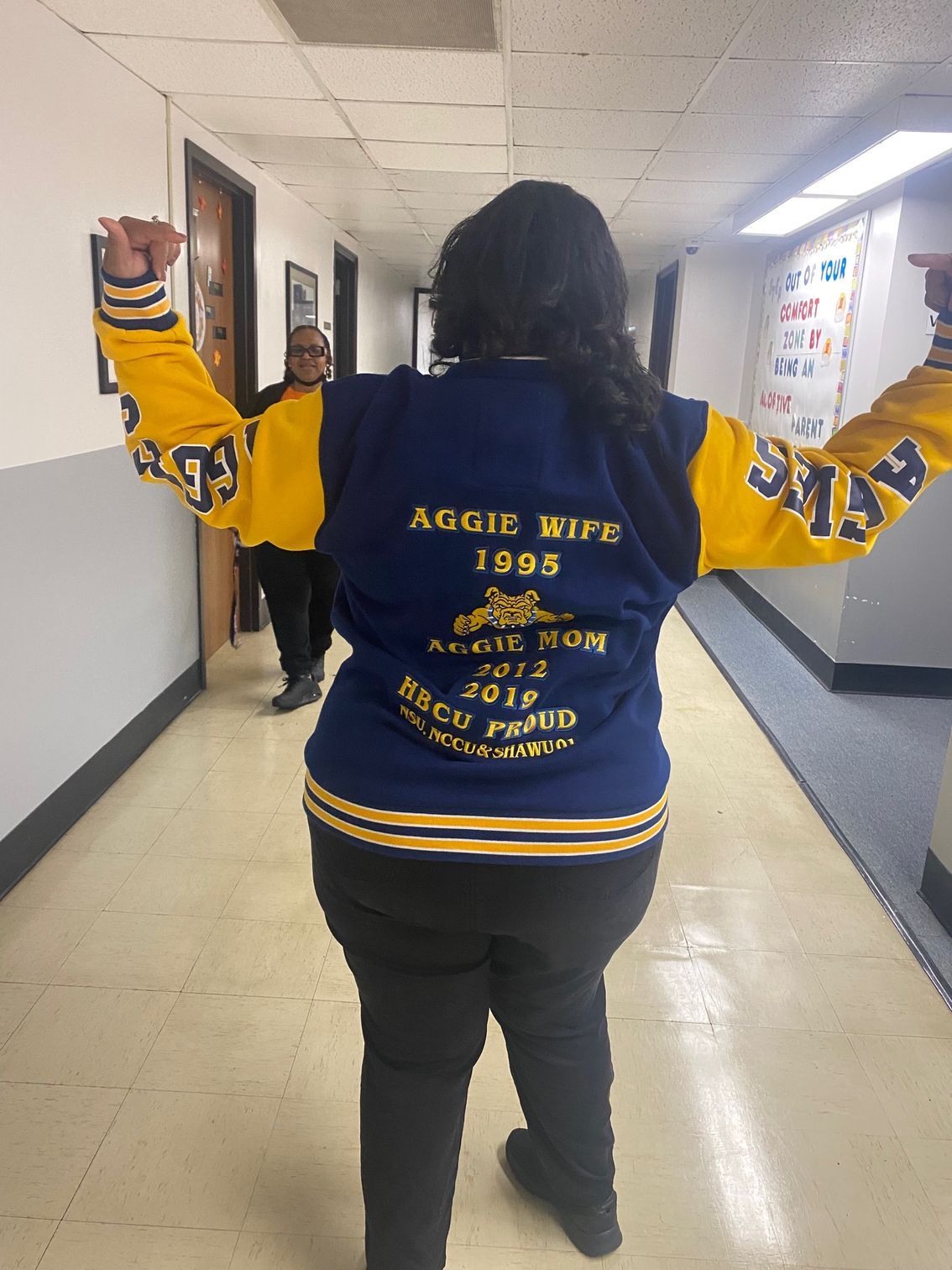 A woman in a blue and yellow jacket is standing in a hallway with her arms outstretched.