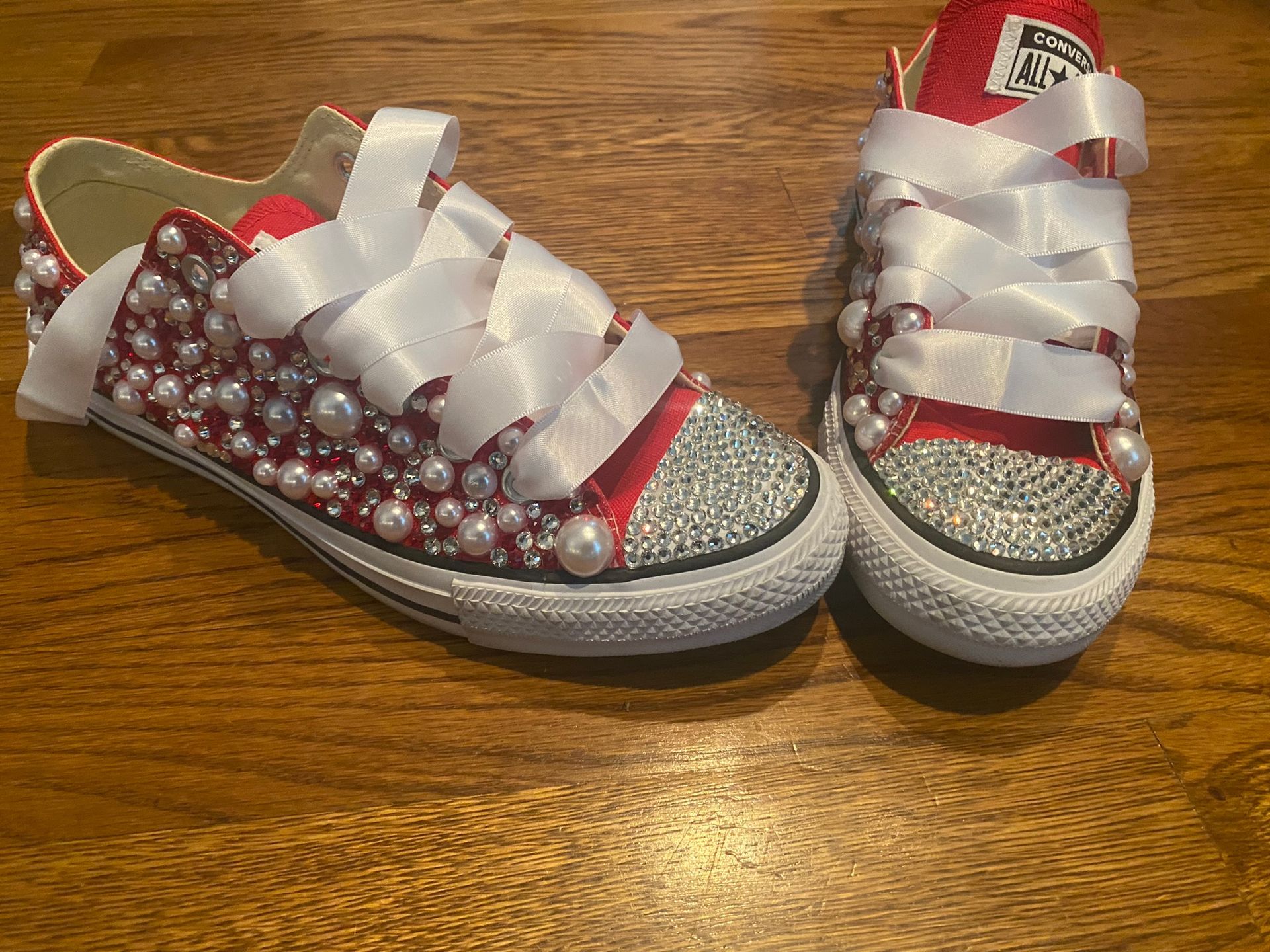 A pair of red and white converse shoes with pearls and ribbons on a wooden table.