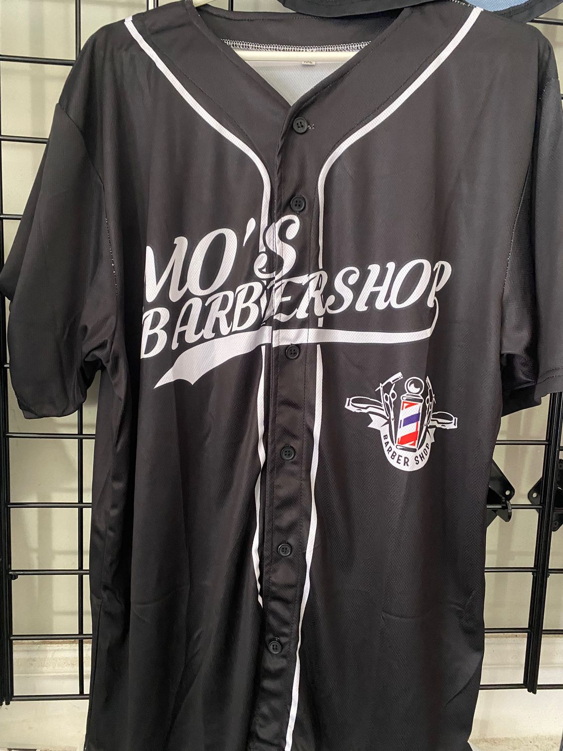 A black barber shop baseball jersey is hanging on a rack.