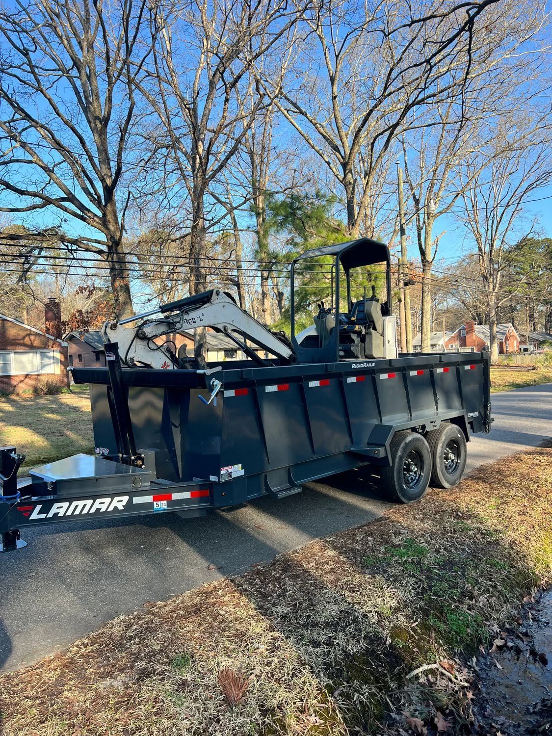 Black Lamar dump trailer parked on a tree-lined street, loaded with construction debris.