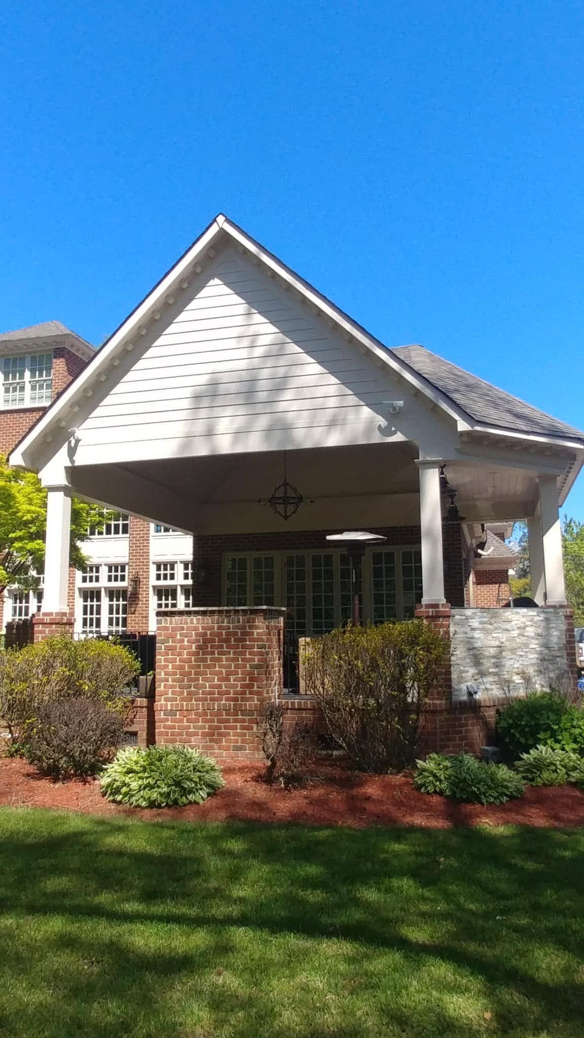 Front porch of a white house with brick columns, red landscaping, and a bright blue sky.