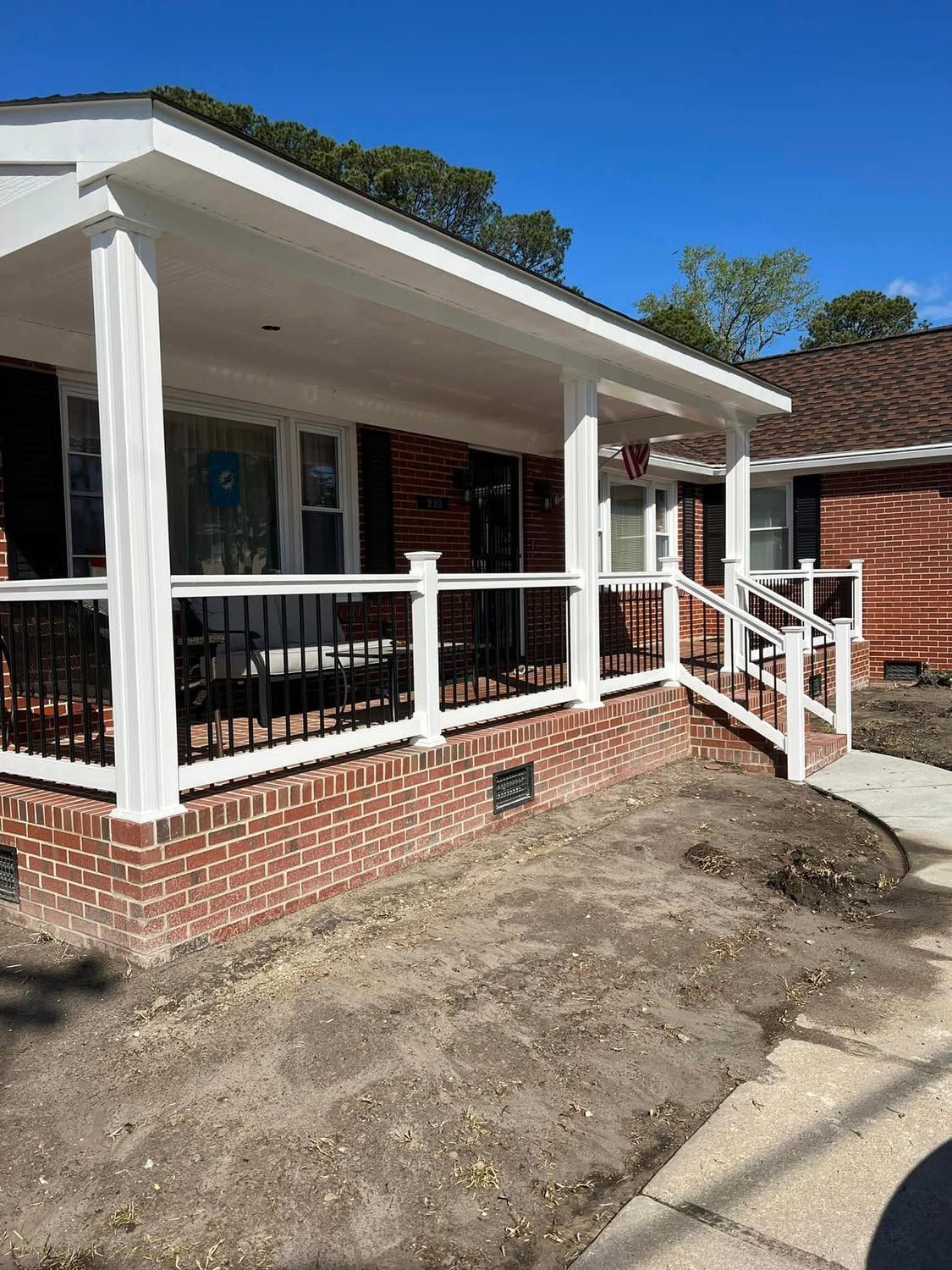Front porch of a brick house with white columns and railing, beside a dirt driveway in sunlight